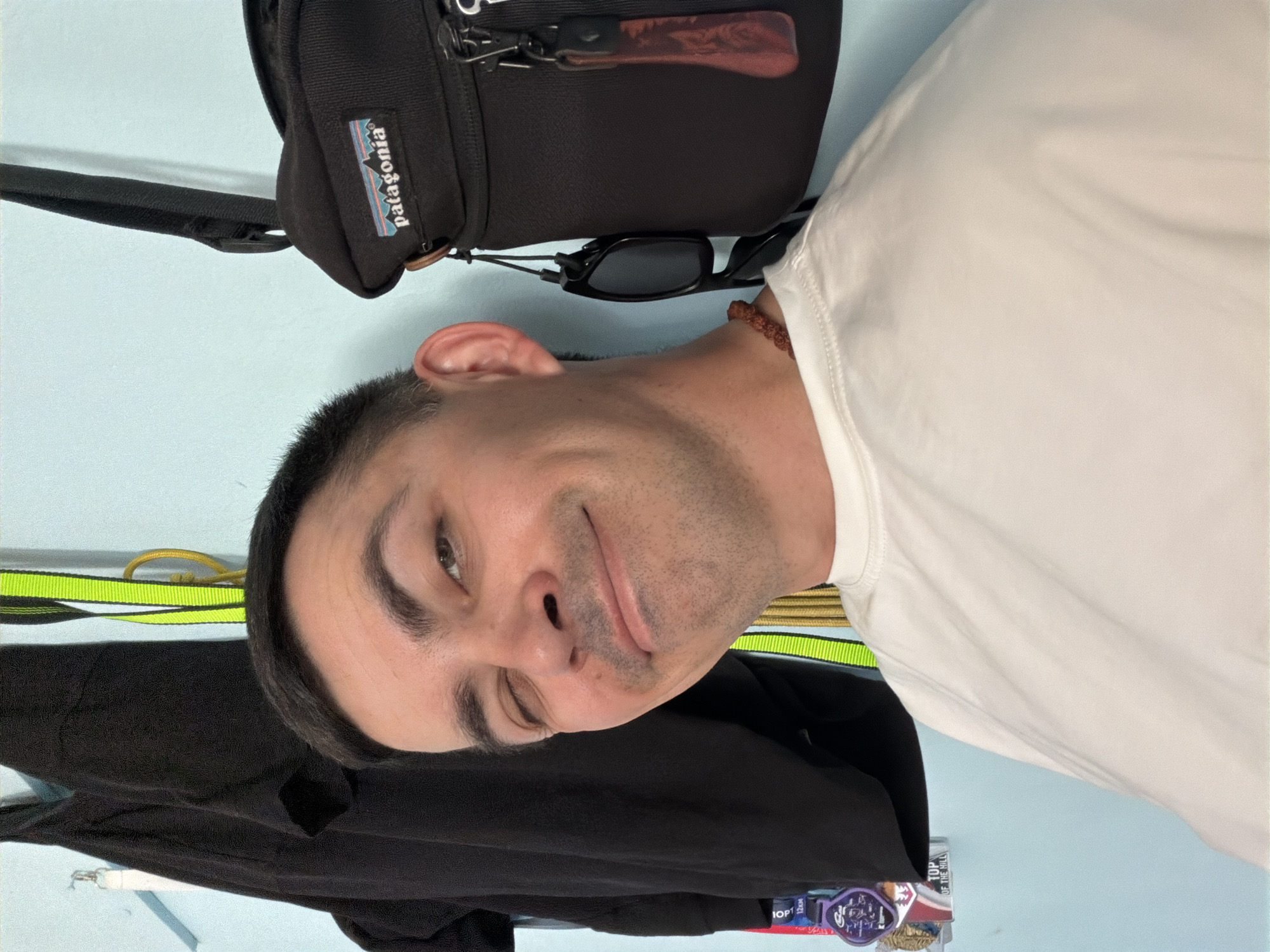 A man is pictured indoors, partially facing the camera with a slight smile. He is wearing a white t-shirt and appears to have a dark-colored bag with a "Patagonia" logo hanging behind him. A pair of sunglasses is draped from the bag. A black piece of clothing hangs in the lower left corner, and yellow and green straps are visible. There is also a small, purple and white sticker or label with text on the lower edge. The background is a light blue-green wall. There is no information in the image to suggest the location is Izhevsk, Russia.
