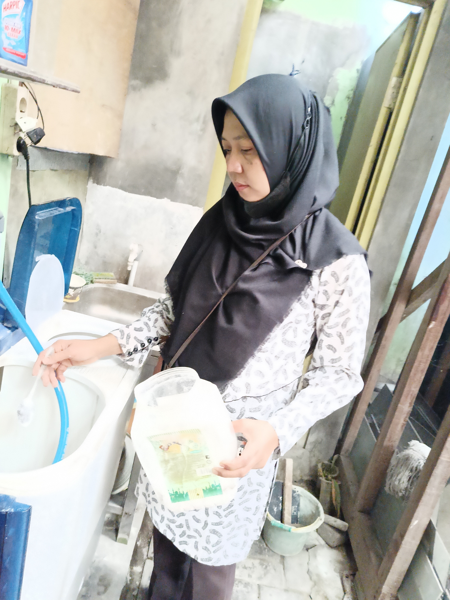 In the city of Sukorini, Indonesia, a woman wearing a black hijab and a patterned shirt stands in what appears to be a laundry area. She is holding a blue plastic hose with a white brush attached, which is inserted into a white washing machine. In her right hand, she holds a large, translucent plastic container filled with a white powdery substance, possibly detergent. The washing machine is white with a blue lid. Behind the woman, a sink is visible, with a faucet. The background shows a concrete wall, a wooden structure with vertical planks, and a mop. A blue bottle labeled "HARPIC 10 MAX CLEAN" is mounted on the wall to the left. The overall environment suggests a domestic or utility space within Sukorini.