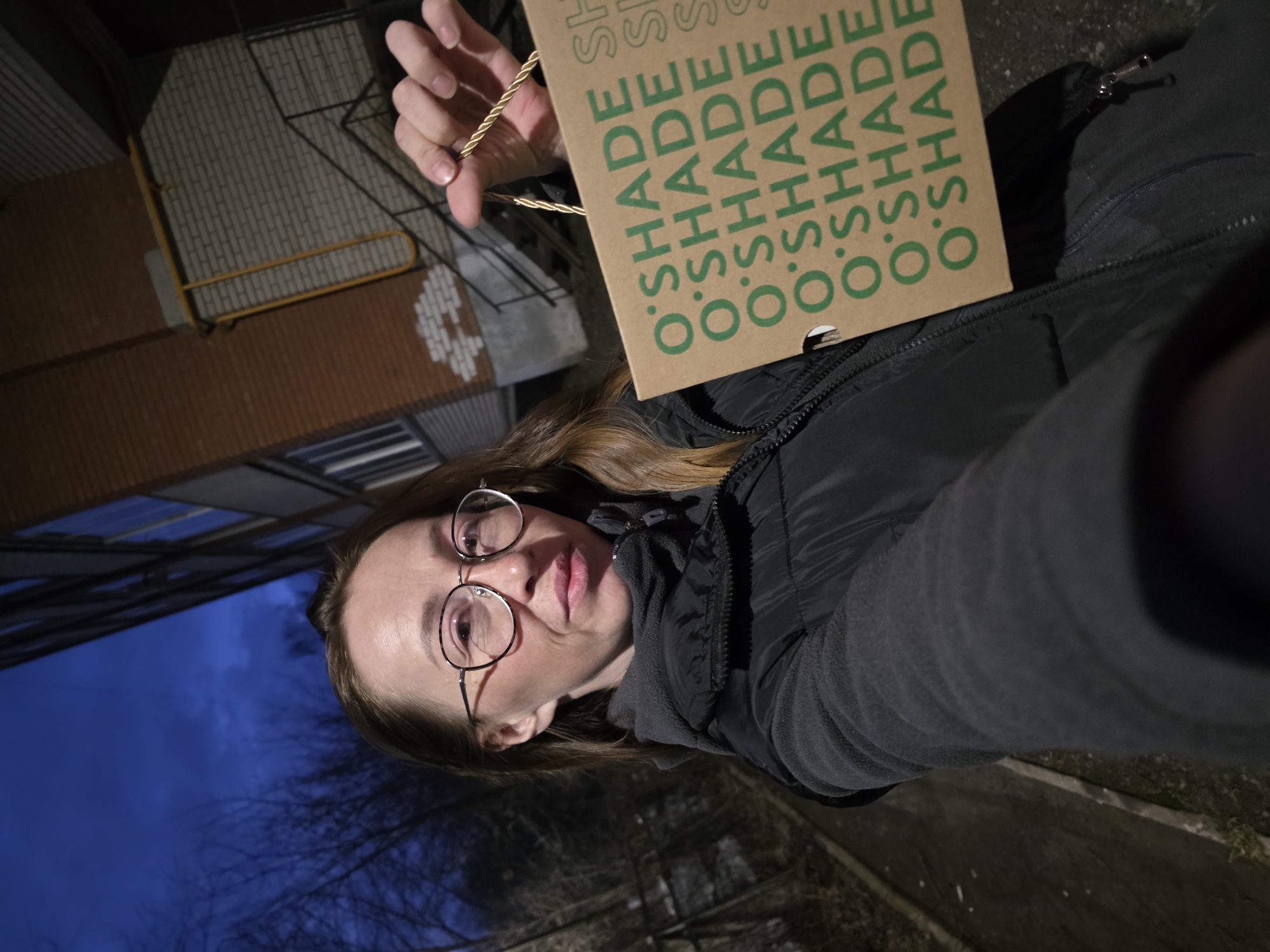 A woman in glasses and a black puffer jacket is holding up a brown paper bag with "O'SHADE" printed repeatedly in green lettering. She is facing the camera with a tilted perspective, suggestive of a selfie. Behind her, the environment suggests an urban setting. A brick building with white accents is visible, along with trees silhouetted against a dusk or night sky. The ground appears to be pavement.
Based on the presence of the "O'SHADE" bag, this scene likely takes place in Chaikovsky, Russia, where the O'SHADE store is located. The exact street or location within Chaikovsky is not determinable from the image.