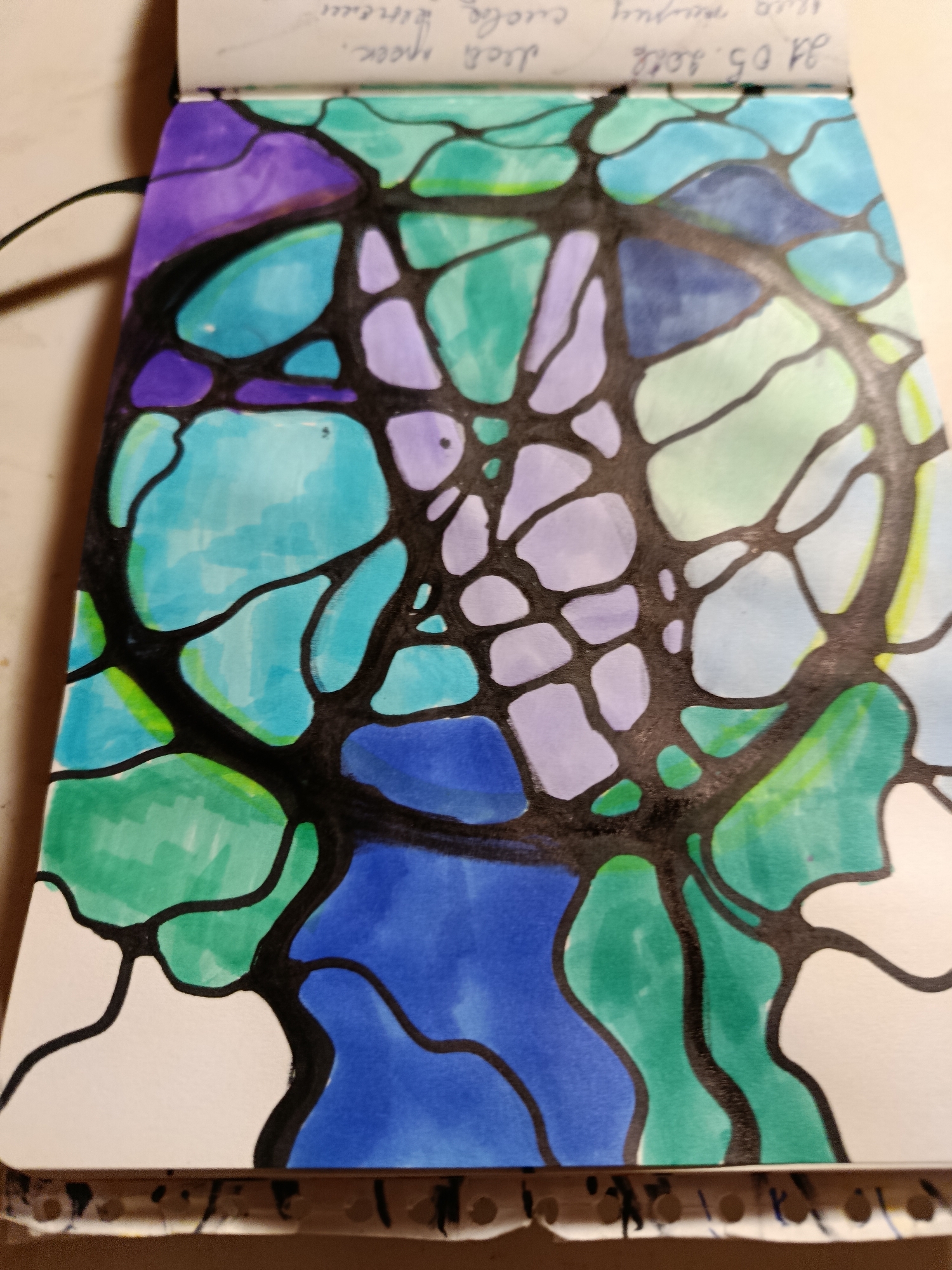 The image shows a close-up view of a notebook open to a page with a colorful abstract drawing. The drawing is done with thick black lines forming irregular shapes that are filled with various shades of blue, purple, and green. Some areas are left white. The notebook itself appears to be spiral-bound, with the edge of the page showing punched holes. Faint handwritten text is visible at the top of the page, partially obscured by the drawing. The background of the image is out of focus, showing a neutral surface. There are no people or discernible city elements in the image.