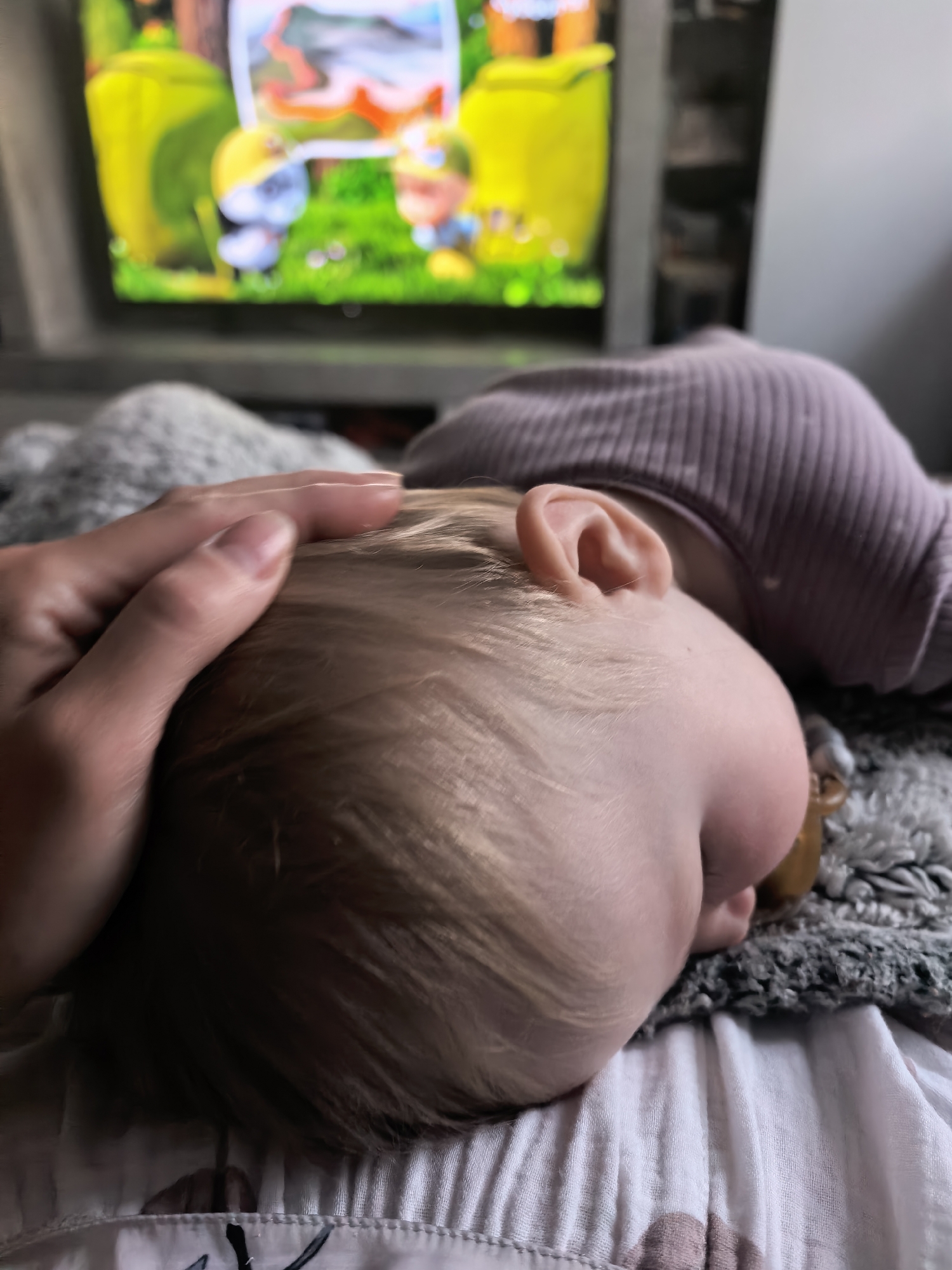 A close-up shot captures a person's hand gently resting on the blonde hair of a sleeping baby. The baby is lying on its side, facing away from the camera, with its head resting on a textured blanket. A pacifier is visible near the baby's mouth. The baby is wearing a purple, ribbed garment. In the background, a television screen displays a colorful, animated scene with cartoon characters and greenery. The overall setting appears to be a home environment. This image does not contain any information that would allow for determination of its location in the city of Sterlitamak, Russia.