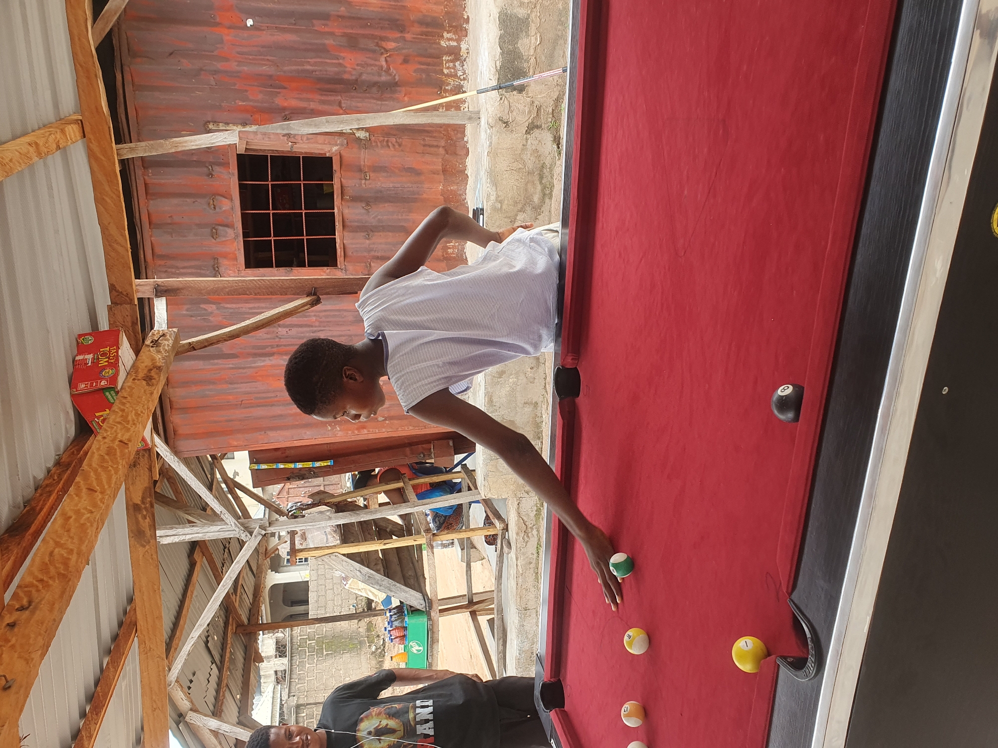 In Lokoja, Nigeria, two young males are engaged in a game of pool at an outdoor venue. One male, wearing a black t-shirt with a lion graphic, stands to the left, observing the game. The other male, in a white sleeveless t-shirt, is actively playing, leaning over the red pool table and in the process of striking a billiard ball with a cue. Several billiard balls are scattered across the table, some numbered. The environment appears to be an open-air establishment with a corrugated metal wall and a wooden roof structure. A pool cue leans against the metal wall. The ground beneath the table is concrete.
