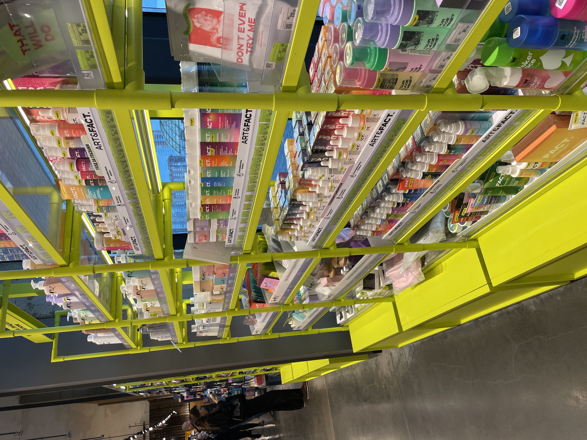 The image shows the interior of a store, possibly a cosmetics or personal care shop, in Krasnoyarsk, Russia. The store features bright yellow shelving displaying various "Art & Fact" branded products. These include items like face creams, serums, and eye care products with Cyrillic labels. The shelves are well-stocked and organized. Several people can be seen at the bottom of the frame, appearing to be shopping and using their phones. The store has a modern and somewhat industrial feel with a concrete floor. Light fixtures hang from the ceiling.