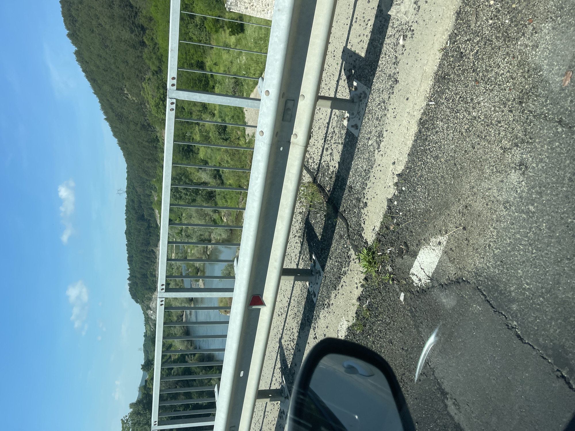 The image, taken from the perspective of a vehicle, captures a scenic view from a road or bridge overlooking a river valley in the area of Balabanovo, Bulgaria. In the bottom right foreground, a portion of a car's side mirror is visible, suggesting an ongoing journey. The immediate right of the frame shows an asphalt road surface meeting a concrete shoulder, both bathed in bright sunlight which casts sharp shadows from the guardrail.
A prominent metal guardrail with vertical bars extends across the mid-ground, separating the road from the vista beyond. A small, red, triangular reflector is attached to the guardrail. Beyond this barrier, a tranquil river winds through a valley, its banks lush with green vegetation and trees.
In the background, a large, verdant hill or mountain, densely covered in trees, rises majestically under a clear blue sky punctuated by a few wispy white clouds. A power line tower is faintly visible on the distant hillside. The bright sunlight and distinct shadows suggest it is daytime, likely late morning or early afternoon, with clear weather conditions. No specific text is visible in the image.
