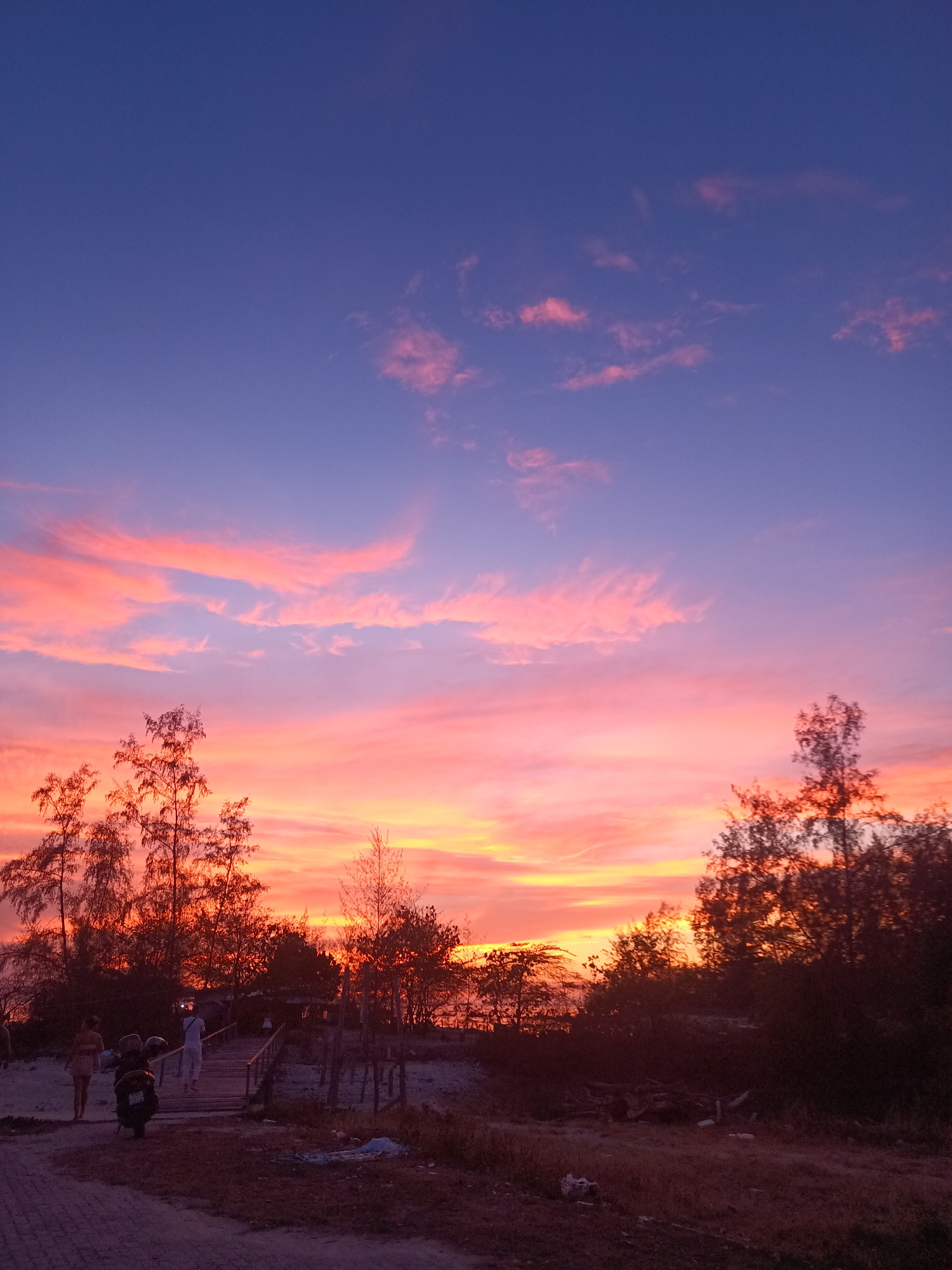 The image depicts a sunset in Baan Sri Thanu, Thailand. The sky transitions from a deep blue at the top to vibrant pink and orange hues near the horizon. Light wispy clouds are scattered throughout the sky. In the foreground, there are silhouettes of trees and foliage. A wooden boardwalk spans a sandy area, with figures of people visible: a woman walking along the beach to the left, and a person ascending the stairs of the boardwalk. A motorbike is parked next to the boardwalk. The ground is sandy with patches of vegetation.