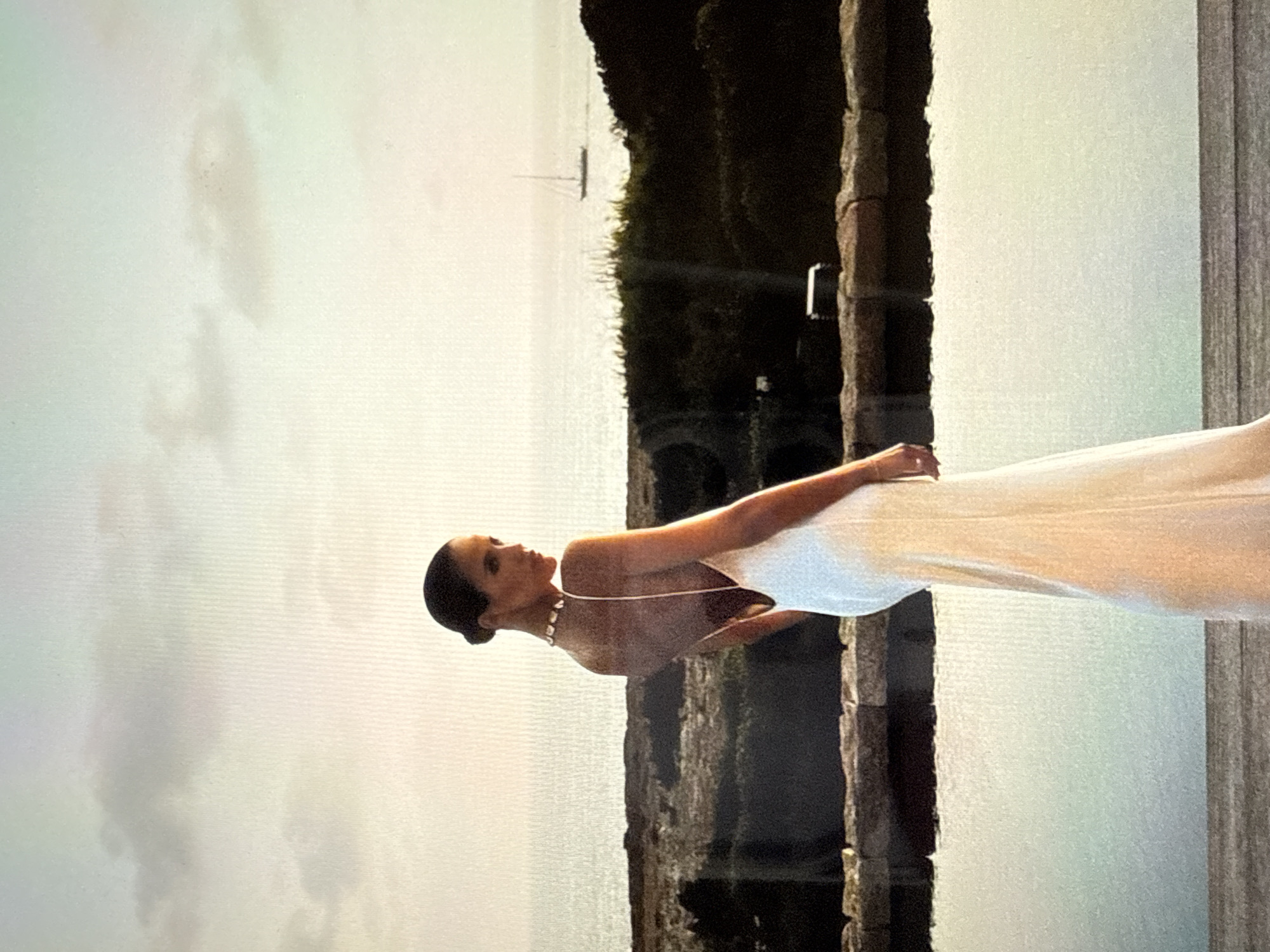 A woman in a white, backless evening gown stands on a wooden deck, looking over her shoulder towards the ocean. A small sailboat is visible on the horizon. The woman's hair is pulled back, and she is wearing a statement necklace. The scene is set at sunset, with soft light illuminating the sky and water. The location is identified as Găvănoasa, Moldova.