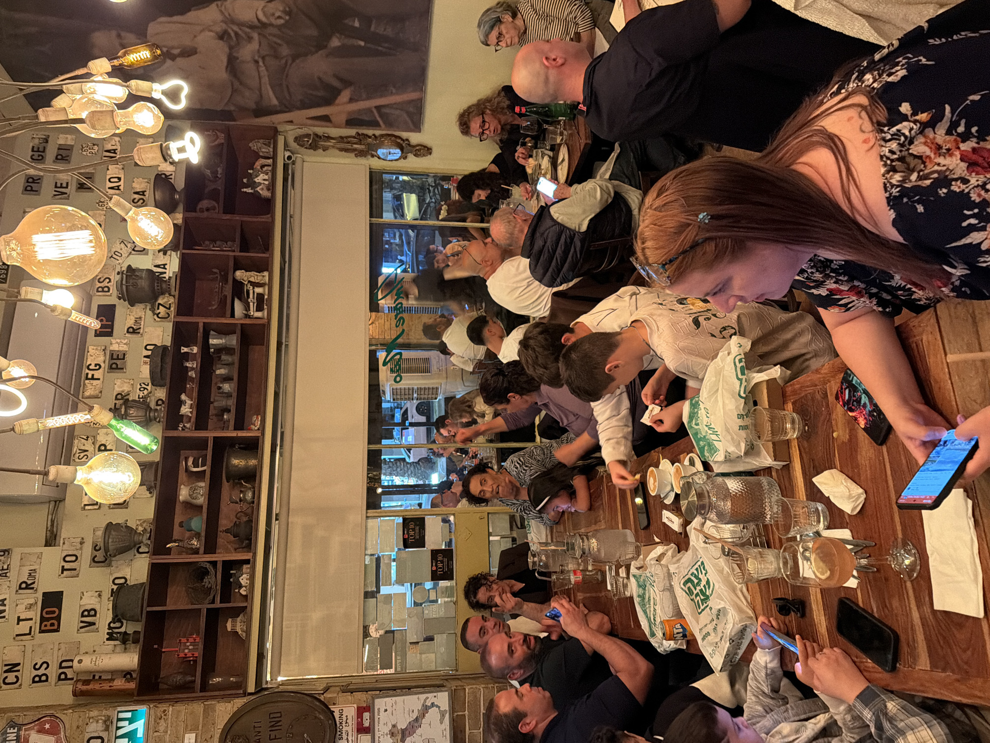 The image depicts a lively indoor dining or social gathering at a restaurant or bar in Tel-Aviv, Israel, likely during the evening. A large group of people of varying ages, from children to adults, are seated at a long wooden table. Many individuals are engaged with their smartphones, while others are seen conversing or observing their surroundings.
The decor is eclectic, featuring a wall adorned with numerous vintage European license plates, including several Italian plates such as "ROMA," "BS" (Brescia), "PD" (Padova), "BO" (Bologna), "AQ" (L'Aquila), "VERI" (Verona), "SA" (Salerno), "CN" (Cuneo), "LT" (Latina), "FG" (Foggia), and "VB" (Verbano-Cusio-Ossola), along with "CZNA" (possibly Czech Republic) and "SV-4". Adjacent shelves display various antique objects like metal pots, vases, and figurines. An industrial-style light fixture with numerous exposed incandescent bulbs, some heart-shaped, illuminates the space.
On the table, there are glasses, carafes of water, coffee cups, and plastic bags with Hebrew text such as "פוצה" (Footsa) and "סות" (Soot), and the name "Imre". Part of the word "ANTA" (likely Fanta) is also visible on one bag. A "No Smoking" sign is clearly visible, displaying "SMOKING" in English and "ممنوع التدخين" in Arabic. Reflections in a window show signs with "TOP 10". People are dressed casually, contributing to a relaxed and social atmosphere.