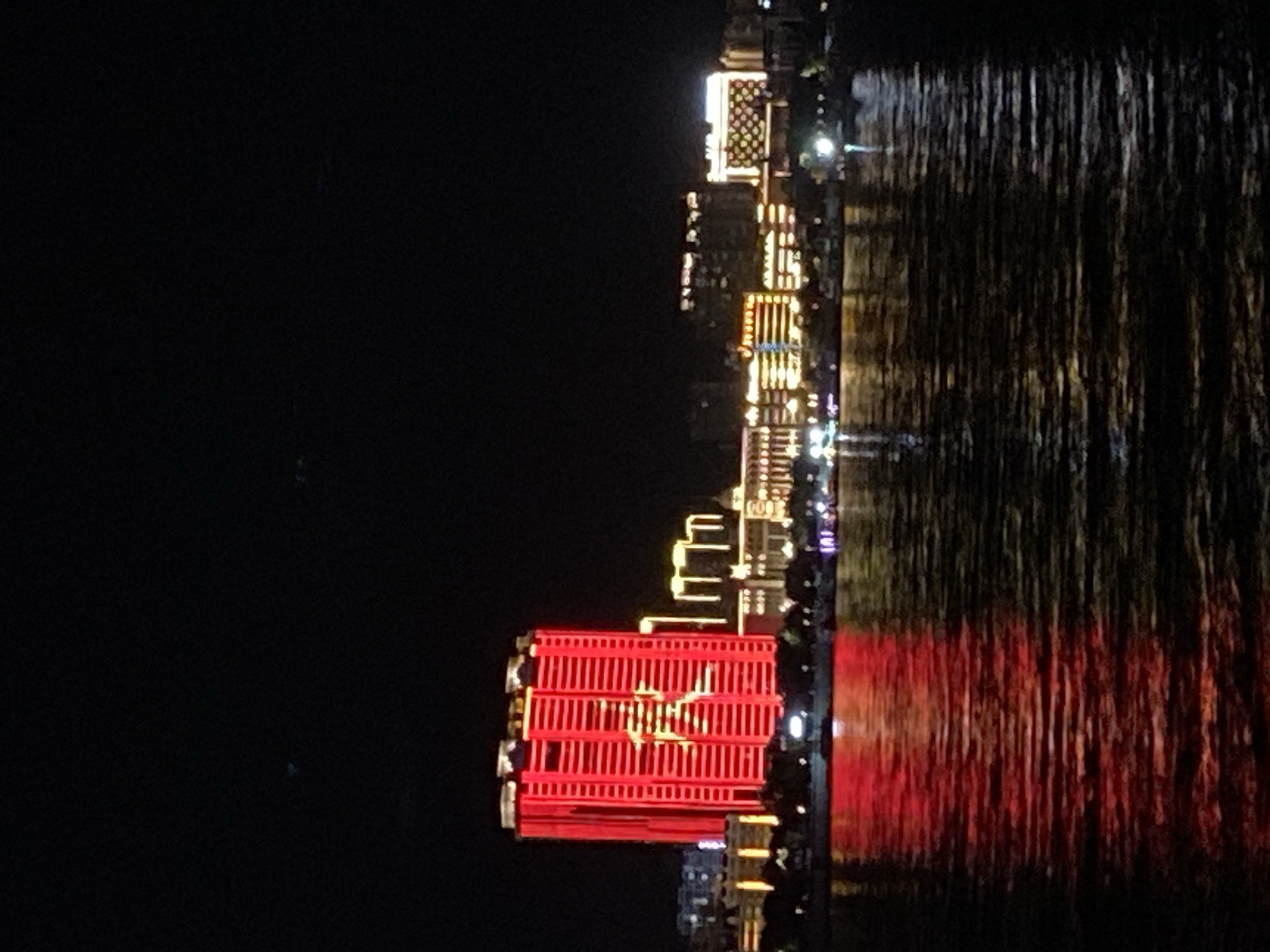This is a nighttime photograph taken from across a body of water, likely the Amur River, looking towards the city of Blagoveshchensk, Russia. The most prominent feature is a tall, rectangular building illuminated in bright red light, with a large, yellow illuminated Chinese character in the center. To the right of this building, other structures are visible, also lit up with yellow and white lights, displaying various patterns and designs. The water in the foreground reflects the red and yellow lights from the buildings, creating streaks of color on its surface. The sky is dark, indicating it is nighttime. The overall scene depicts a vibrant, illuminated cityscape at night.