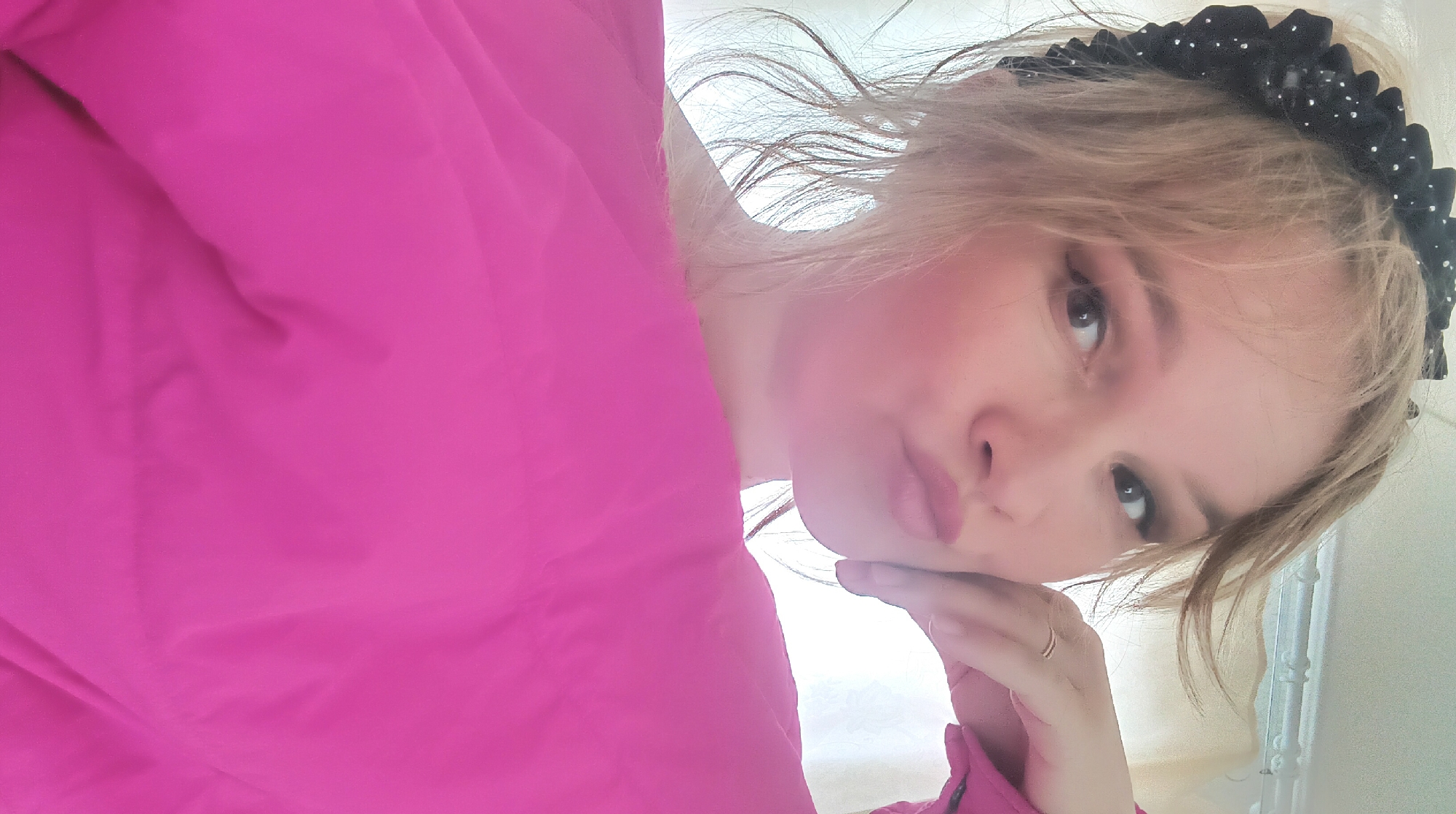 A close-up portrait captured indoors within the city of Ольховка, Russia, depicts an adult female. She has fair skin, blonde hair, and brown eyes directly engaging the camera. A black headband with ruffled fabric and small white embellishments is worn in her hair. Her lips are pursed, exhibiting light pink lipstick, and eye makeup is visible. Her right hand, adorned with a thin gold-colored ring on the ring finger, is positioned with fingers lightly curled under her chin. The lower portion of the frame is dominated by a bright magenta garment, likely a top or jacket. The background is light-colored, out of focus, and indistinct, showing a faint patterned surface in the upper right quadrant.