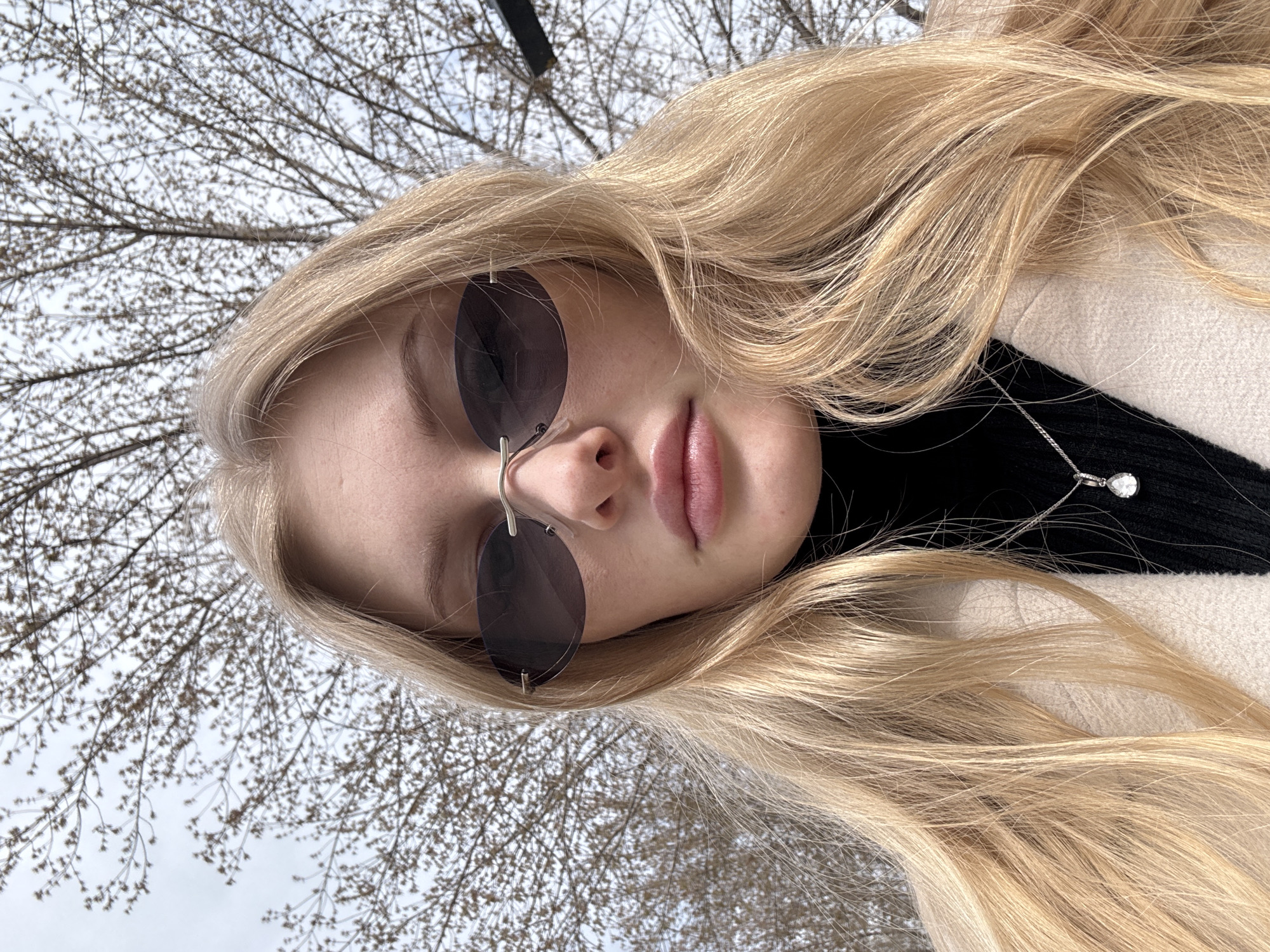 This close-up portrait features a young person, likely female, with long, radiant blonde hair cascading around her. She wears oval-shaped dark sunglasses that cover her eyes, and her lips are slightly parted in a subtle expression. Her attire includes a black top, possibly a turtleneck, visible beneath a light beige or cream-colored coat. A delicate silver chain necklace with a teardrop-shaped, clear pendant rests on her black clothing.
The image is captured from a low vantage point, looking upwards. The background is dominated by a dense network of bare tree branches, indicative of late autumn, winter, or early spring, set against a bright, overcast sky. This outdoor setting suggests a temperate climate, consistent with locations like Perm, Russia, where such tree dormancy is common. The time of day is clearly daytime, characterized by ample, diffused light. There is no visible text in the media.