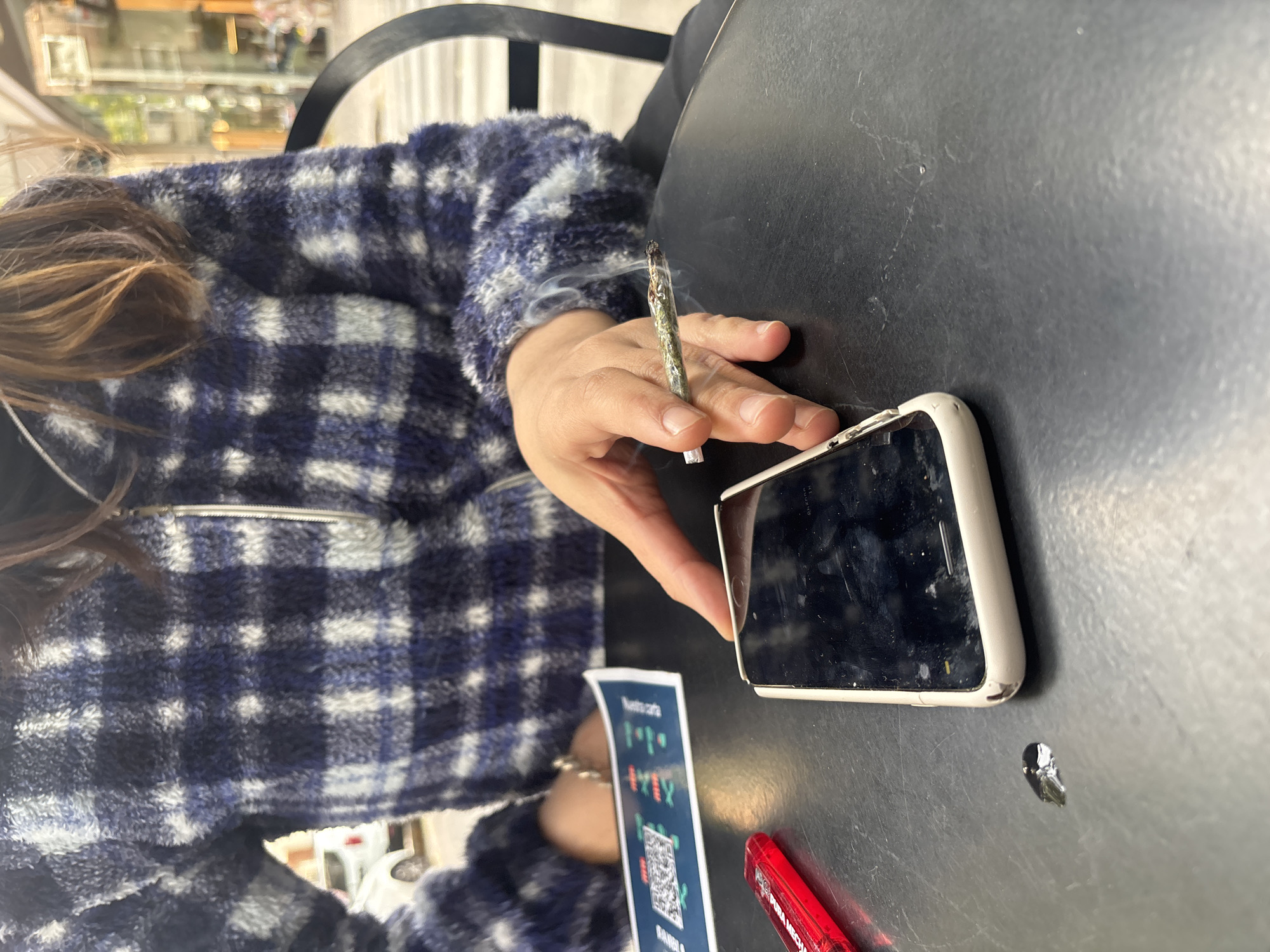 An individual wearing a blue and white checkered fleece jacket sits at a black table outdoors. The person is holding a lit, hand-rolled cigarette (likely marijuana) in their right hand. A white cell phone with a cracked screen lies on the table to the right of the person's hand. A promotional card with blue and white text and a QR code is placed below the phone. A red object is also visible near the edge of the table. The setting suggests a casual outdoor cafe or restaurant. The location is likely Buenos Aires, Argentina, based on the Spanish text on the promotional card.