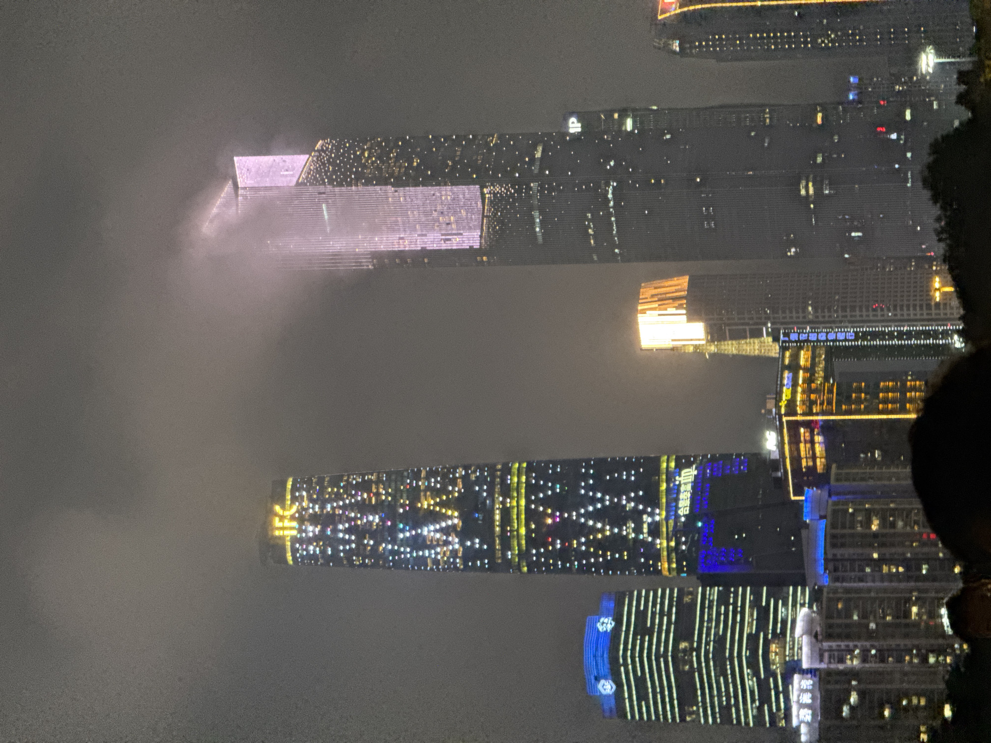 At night in Yuexiu District, China, a cityscape is visible through a dense fog. Illuminated skyscrapers dominate the view. The fog obscures the lower portions of the buildings and creates a sense of the structures floating in the night sky. Building lights shine through, creating a display of colors like gold, blue, and white. On the right edge, a dark silhouette of a person's head is partially visible.