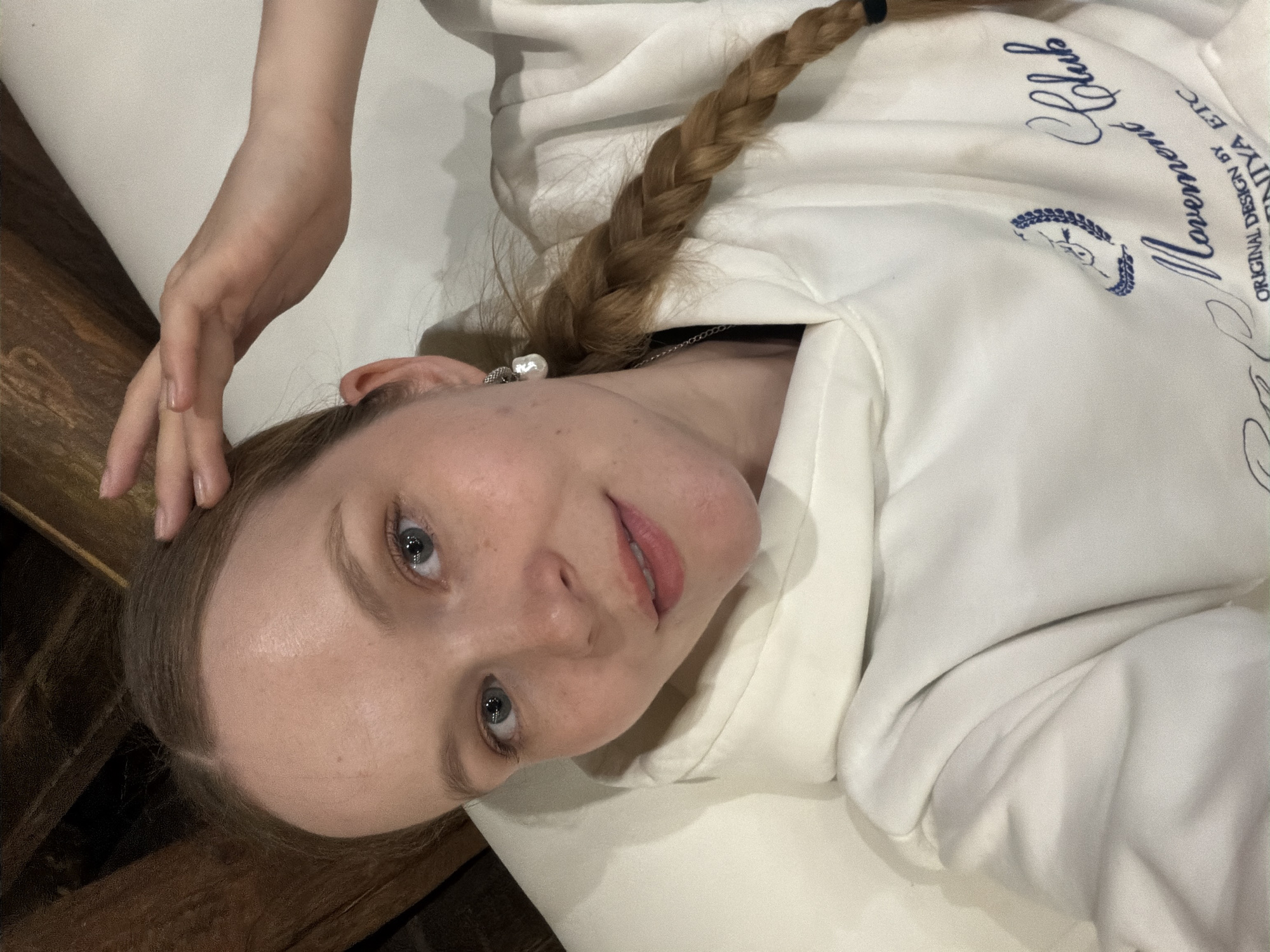 A young person, likely female, with fair skin, light-colored eyes, and light brown hair styled in a braid, is depicted in this indoor portrait. She is lying down, possibly on a bed or couch, against a light-colored surface. A wooden surface is visible in the background, to the left. She is wearing a white hooded sweatshirt with blue text and a small blue emblem on the front. Visible text on the sweatshirt includes words like "Club", what appears to be "Lemon" or "Moment", another cursive word that could be "Moments" or "Mornin'", and the phrase "ORIGINAL DESIGN BY NIYA ETC". The emblem resembles a stylized head within a wreath.
Her right hand is raised and resting near her head. A pearl earring is visible in her right ear, and a thin silver chain is seen around her neck. She looks directly at the camera with a relaxed expression and a slight smile. The image is taken from a slightly overhead perspective. There are no specific visual cues within the image to indicate the time of day or the location of Yus'ki, Russia, other than the provided context.