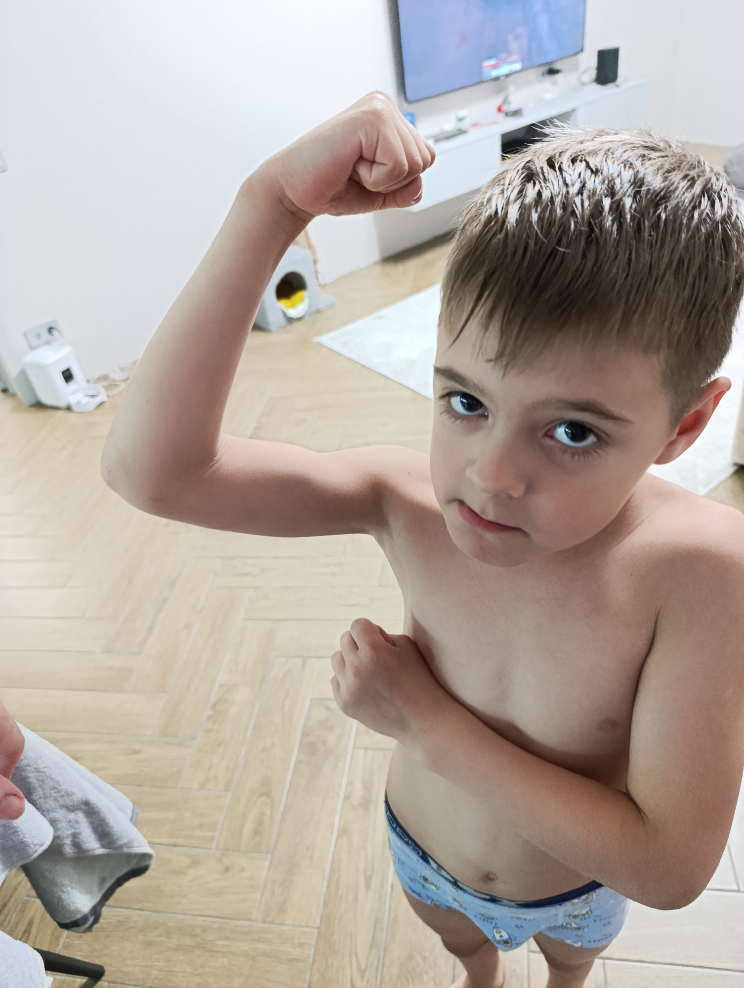A high-angle shot depicts a young boy flexing his left arm, creating a bicep bulge, while his right hand rests on his chest. He is shirtless, wearing patterned blue underwear. His hair is wet. The background shows a modern interior with light-colored flooring, a white cabinet, and a television. There is a white, small appliance and a cat house-like structure in the background. A portion of a gray towel is visible in the lower left corner. There are no indicators that this is in the city of Chaikovsky, Russia.