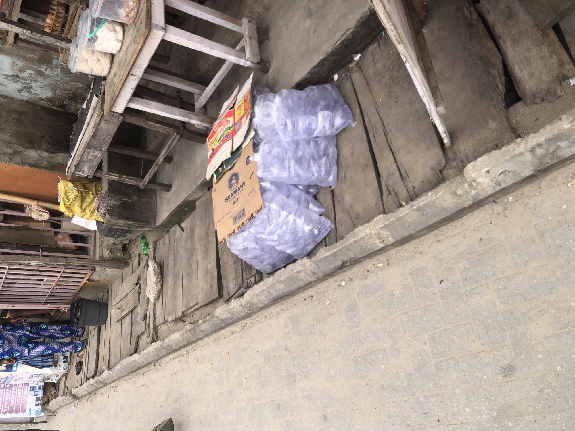 An outdoor scene shows a walkway next to a street in Ado, Nigeria. The walkway is made of interlocking concrete pavers and is slightly sloped. Adjacent to the walkway is a wooden plank structure, possibly a makeshift path or display area. On this wooden structure, several clear plastic bags filled with what appears to be ice are piled up. A cardboard box, possibly used for packaging or as a table, rests on top of some of the ice bags. Further back, a wooden table holds containers of food items, and in the background, a person in a blue and white patterned garment is visible. The overall environment appears to be a street market or a commercial area with signs of daily commerce.