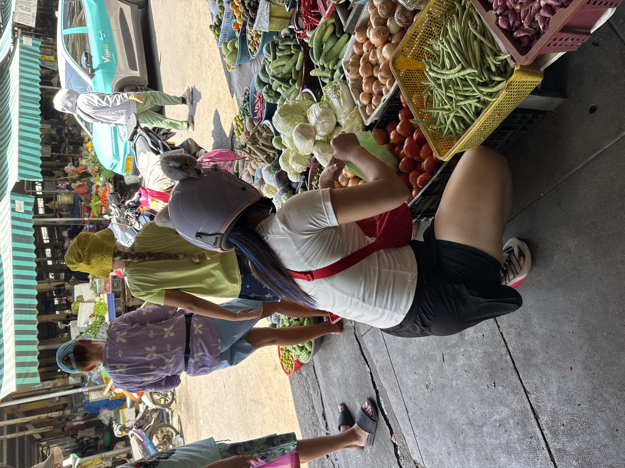 The image captures a bustling outdoor market scene in Nha Trang, Vietnam. In the foreground, a person wearing a white t-shirt, black shorts, and a helmet with ear-like decorations is crouched down, sorting vegetables, likely green beans, into a yellow plastic basket. The person's left arm is extended, holding a green plastic bag. To the left, two children are standing, one in a light purple patterned shirt and blue shorts, and the other in a green t-shirt and dark blue shorts. Behind them, a variety of fresh produce, including tomatoes, onions, cabbage, cucumbers, and peppers, are displayed in plastic crates and baskets. In the background, a turquoise and silver car is parked, and market stalls with striped awnings are visible, suggesting a lively commercial area. The ground is paved with concrete, showing cracks and some debris. The lighting indicates a sunny day, casting shadows and highlighting the vibrant colors of the market goods.