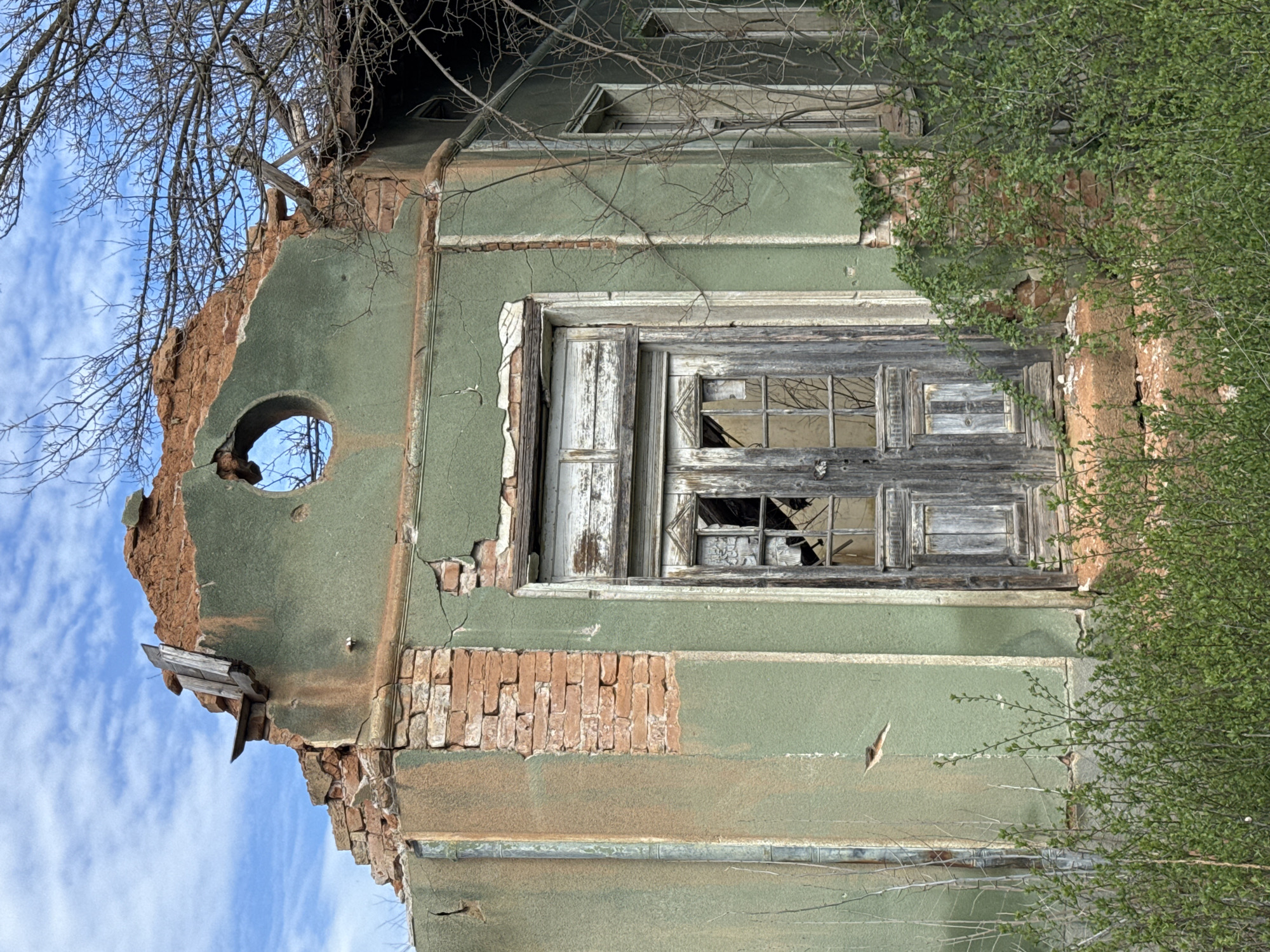 The image shows a derelict building in Krushovitsa, Bulgaria. The building's facade is pale green with exposed brickwork in areas where the plaster has deteriorated. A broken window with a deteriorated wooden frame is visible. Part of the building's upper section is missing, revealing a hole in the wall. Bare tree branches are seen against a blue sky with white clouds in the background. A green, bushy plant is growing along the side of the building.
