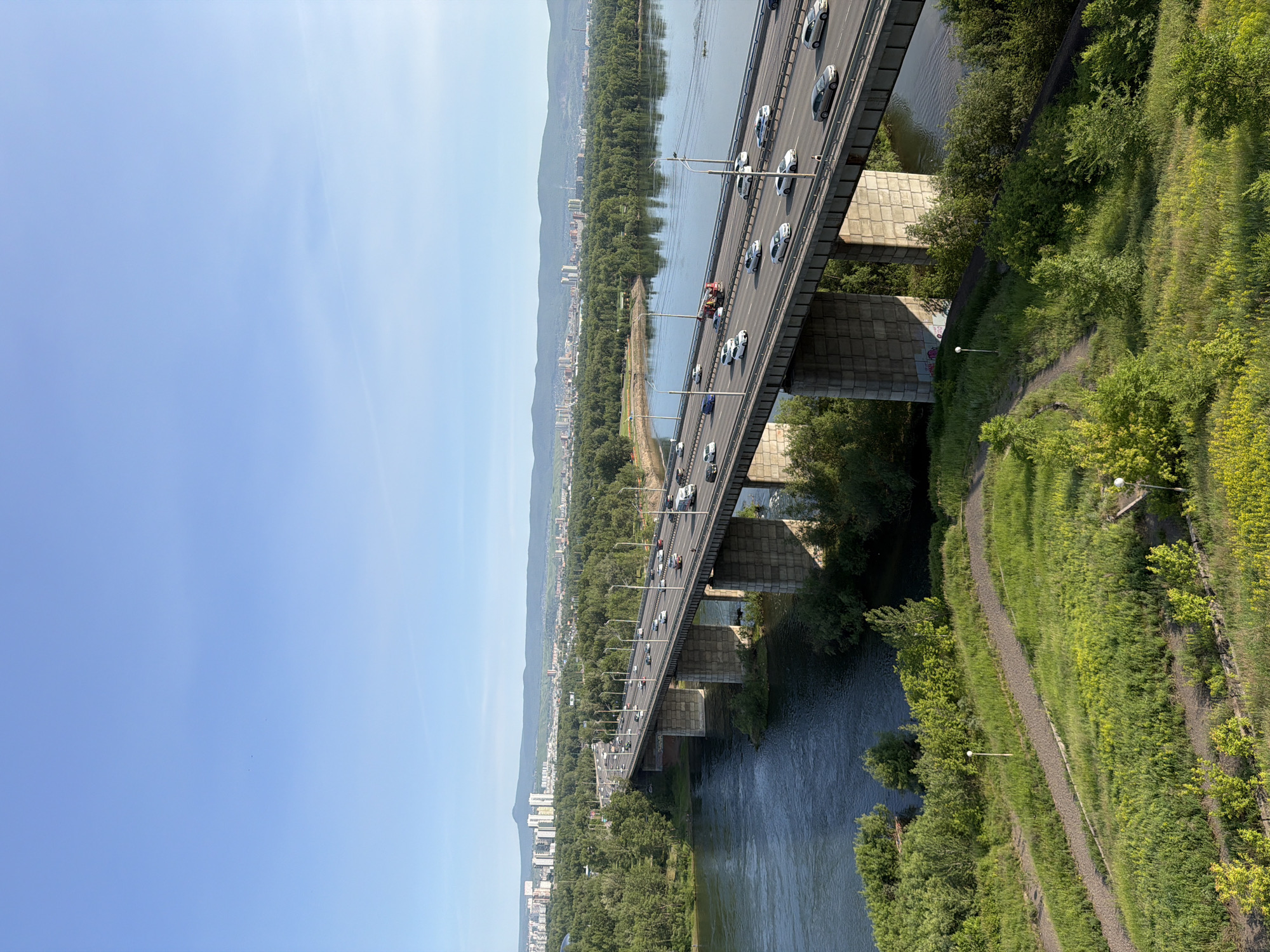The image captures a scenic view of a bridge spanning a wide river, with the city of Krasnoyarsk, Russia, visible in the background. The bridge is a prominent subject, carrying a steady flow of traffic, including cars and possibly trucks. The river below appears calm, reflecting the clear blue sky and the lush green trees that line its banks.
The setting is outdoors on a bright, clear day, suggesting it is daytime, likely late morning or afternoon, given the quality of light. The abundance of green foliage indicates a warm season, possibly summer. The distant cityscape, with its buildings and mountains, provides a sense of the location. There is a walking path visible on the right side of the image, bordered by green grass and trees, suggesting the area is used for recreation.
Notable details include the concrete pillars supporting the bridge, the streetlights lining the roadway, and the variety of vehicles in motion. The image is taken from an elevated perspective, likely from another bridge or a high vantage point, offering a panoramic view of the urban landscape and the natural environment. No text is visible in the image.