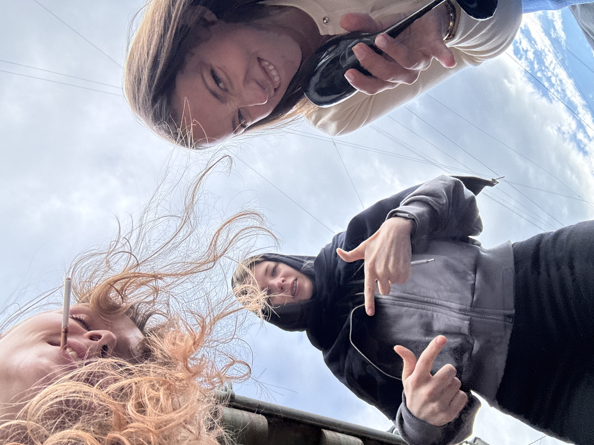 The image shows a low-angle, wide shot of three individuals looking down at the camera against a backdrop of a cloudy sky with power lines visible. The person on the far left has reddish-brown hair and is positioned upside down, with their mouth open and a cigarette between their lips. In the center, a person wearing a black hoodie and headscarf points at the camera with both hands, their expression neutral. To the right, a woman with long brown hair smiles broadly, holding a black object, possibly a hairbrush or a similar item. The scene appears to be outdoors, with no specific landmarks indicating Saint Petersburg, Russia. The lighting suggests daytime.