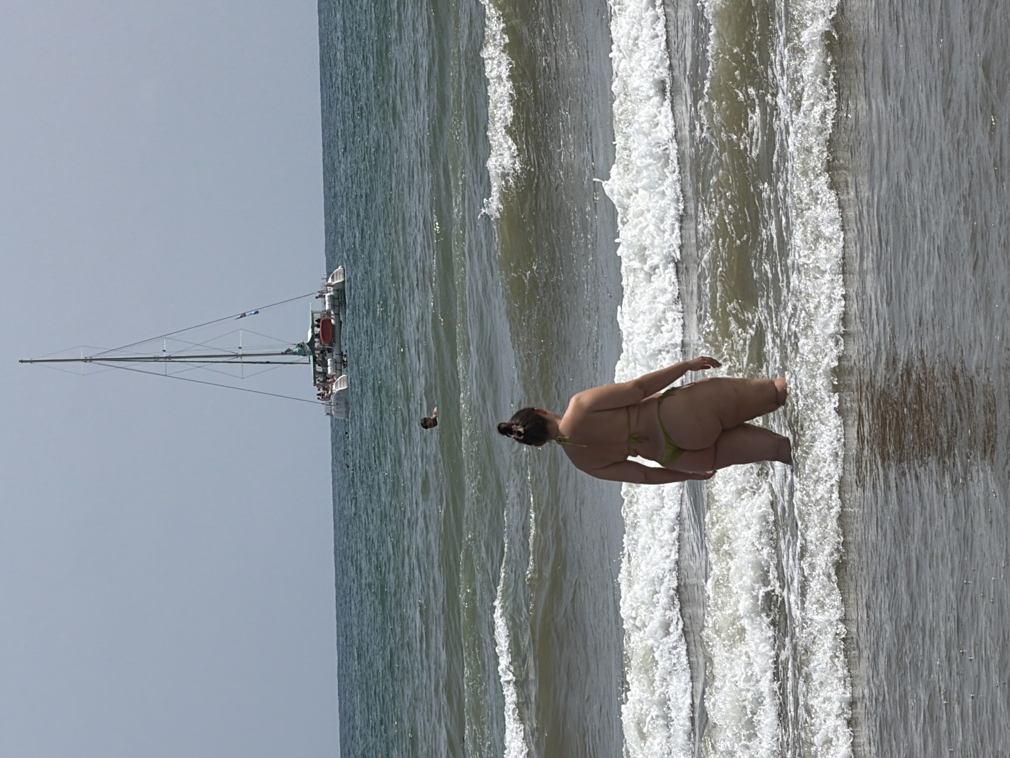 A person in a green bikini stands in the ocean with waves breaking around their legs. The person is facing away from the camera. In the background, a catamaran boat with several people on deck sails on the water under a clear sky. The ocean is choppy with white-capped waves.