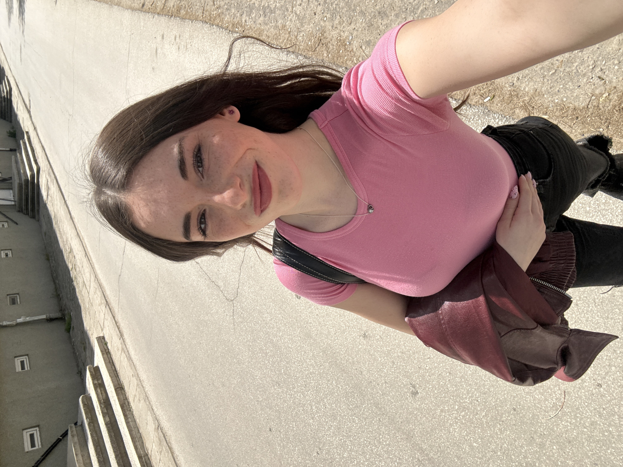 A young woman is captured in a sunlit outdoor selfie, smiling gently towards the camera. She has long, dark brown hair and light-colored eyes, with visible freckles across her nose and cheeks. She is wearing a fitted, ribbed pink short-sleeved t-shirt and black pants. A delicate silver chain necklace with a small heart-shaped pendant adorns her neck, and she has a tiny pink stud earring in her right ear. Her nails are manicured with pink polish decorated with small white specks. A black shoulder bag with white stitching is visible over her right shoulder, and a maroon or dark red jacket is draped over her left arm.
The background shows a paved surface, likely a street or sidewalk, bathed in bright sunshine. To the left side of the frame, a light-colored building with multiple small, rectangular windows and a set of concrete steps are visible. The strong shadows suggest a clear, sunny day, indicating daytime hours. The architectural style hints at a European urban setting, consistent with the location context of Plovdiv, Bulgaria. The woman's expression conveys a pleasant and relaxed mood. No visible text is present in the image.