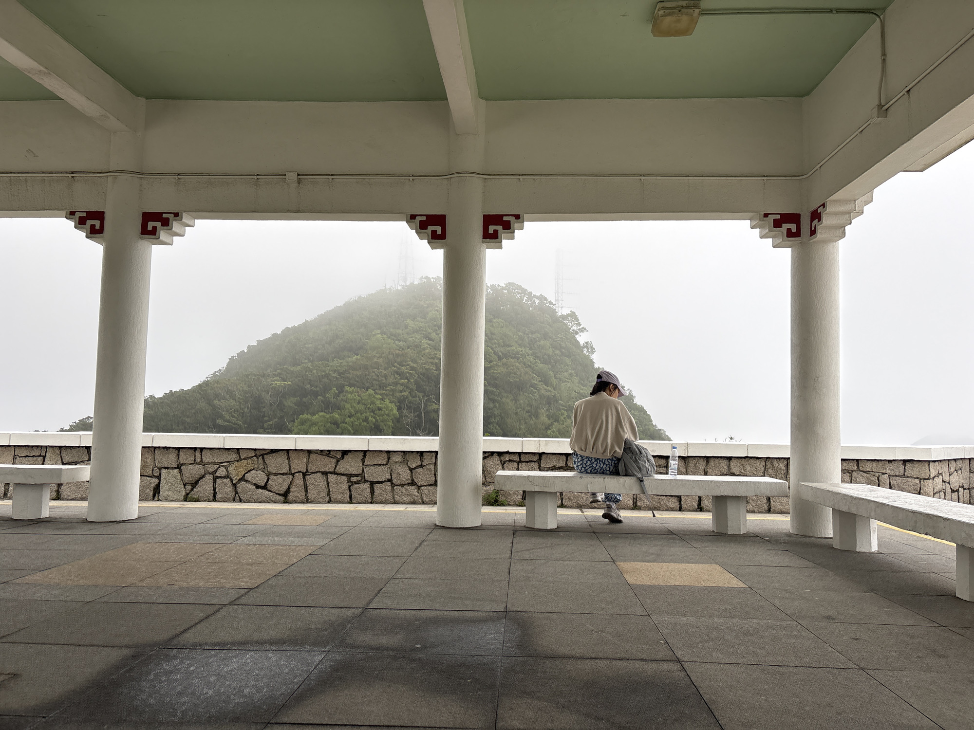 A medium shot depicts a person sitting on a white bench in a covered shelter with a view of a mountain shrouded in mist. The person is facing away from the viewer and is wearing a light-colored jacket, patterned pants, and a cap. A gray backpack and a water bottle are placed on the bench next to them. The shelter has white columns with red ornamental details at the top and a green ceiling. The floor is made of square tiles in varying shades of gray and brown. Outside the shelter, there is a low stone wall and a view of a green, tree-covered mountain obscured by fog. Transmission towers are faintly visible on the mountain's peak. The scene is set in Hong Kong, China.