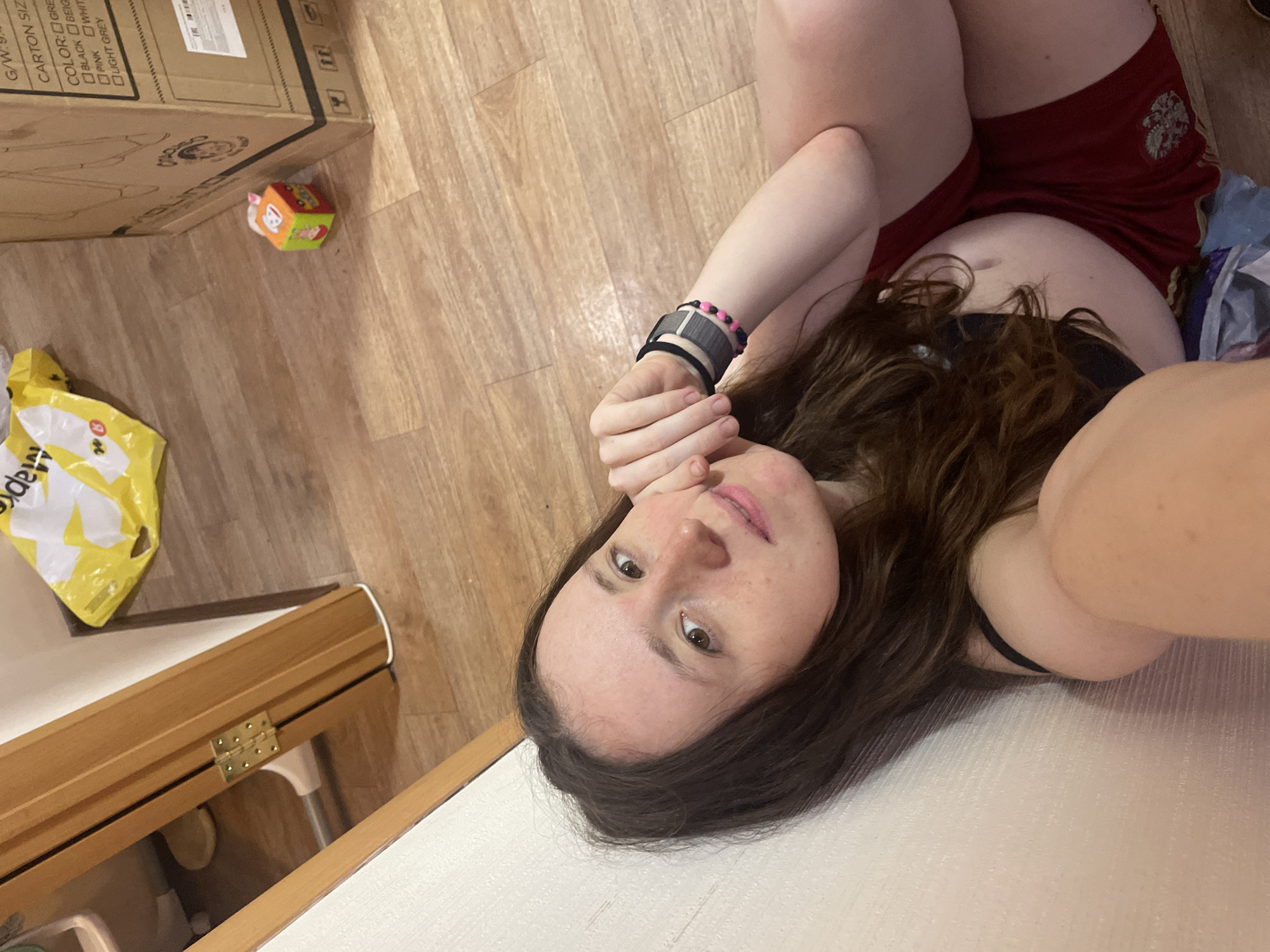 A person with long brown hair, wearing a black top and red shorts, is lying on a light-colored surface, possibly a bed or sofa. The person is looking at the camera with their hand near their face.
In the background, there is a wooden floor, a cardboard box with some printed text (including the words "CARTON SIZE" and color options), a yellow plastic bag, and a child's toy cube. The scene appears to be indoors.
There is no indication of the location being in Usol'e, Russia.