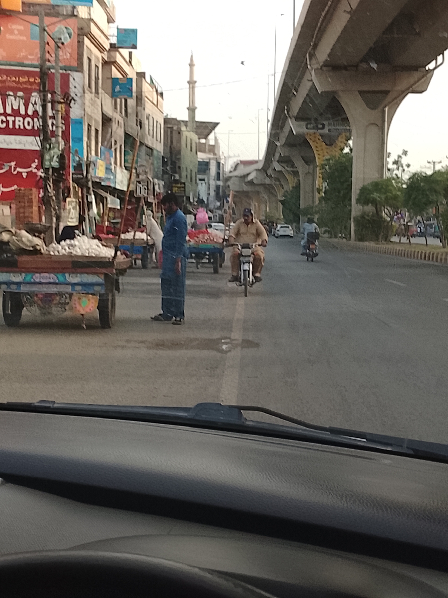 A street scene in Gulgasht Colony, Pakistan, depicts a road occupied by pedestrians, carts laden with produce, and motorcyclists. An elevated highway structure dominates the right side of the frame. The foreground features a car dashboard from which the photo is taken. In the middle ground, a man in a blue shalwar kameez stands near a cart, while another man rides a motorcycle towards the camera. Further back, a second motorcyclist follows, with additional pedestrians and vehicles visible on the right. Buildings line the left side of the street, interspersed with shops. A minaret rises in the distance.