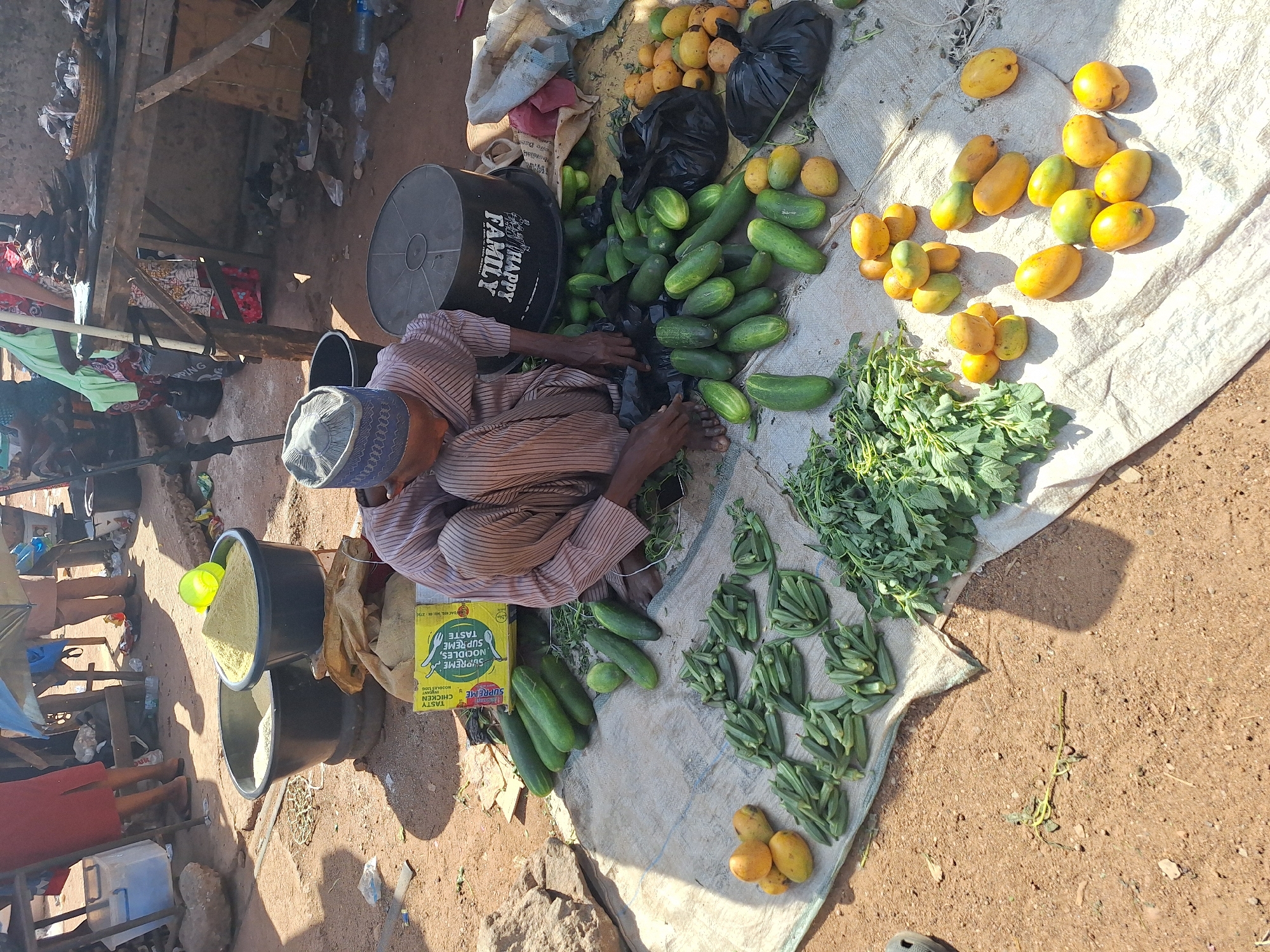 The image depicts a vendor at an outdoor market in Kakuri Gwari, Nigeria. The vendor, an older man wearing a traditional cap and striped brown clothing, is seated on the ground next to a black container with the words "HAPPY FAMILY" printed on it. Spread out on a white cloth in front of him are various fruits and vegetables, including cucumbers, mangoes, leafy greens, and okra. A box of "Supreme" brand chicken noodles is positioned near the cucumbers. Two stacked black buckets, one containing a yellow substance, are also nearby. The ground is reddish-brown dirt and the setting appears to be a bustling market area with other people and stalls visible in the background.