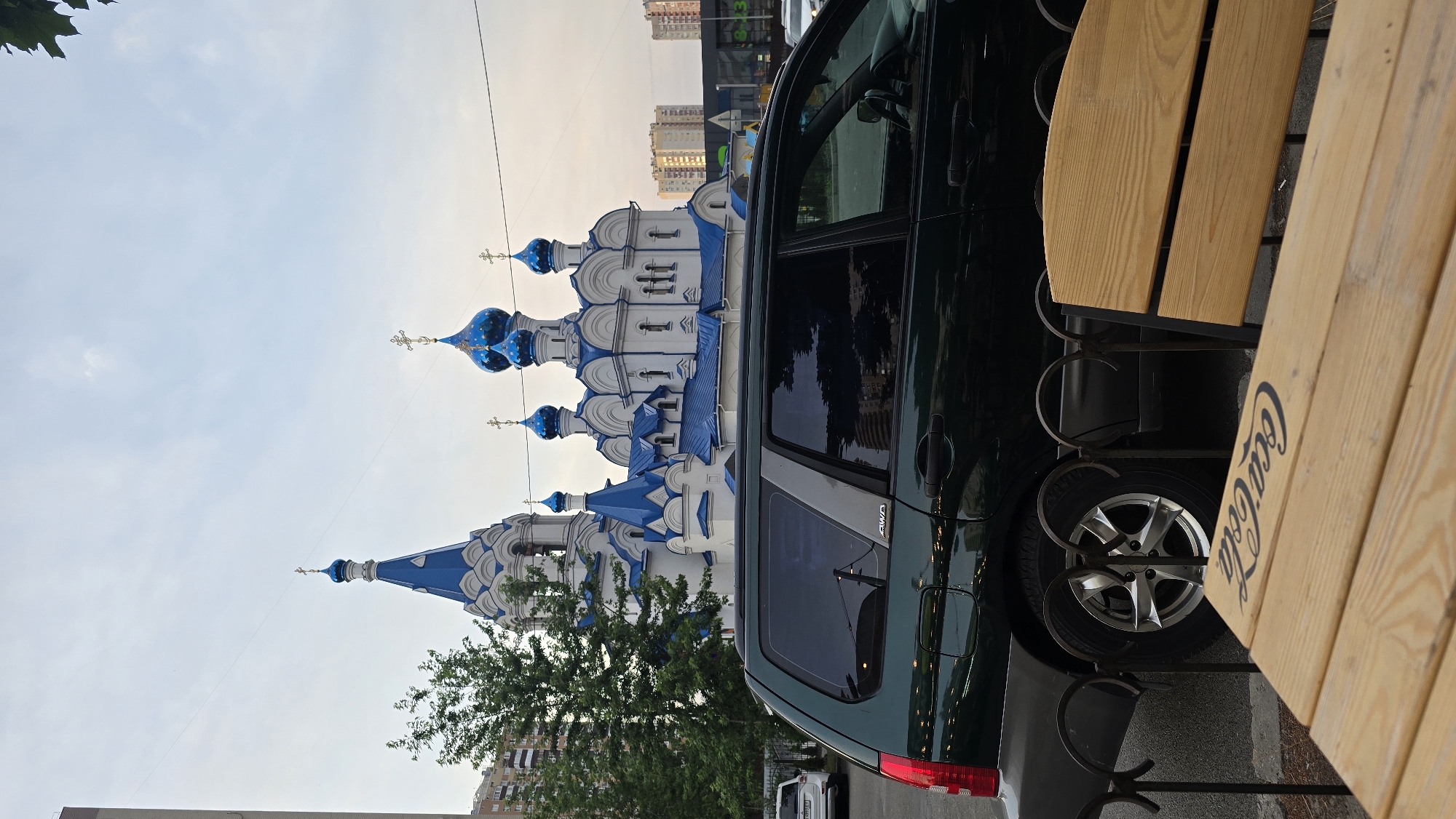 A dark green SUV is parked in front of a white church with blue domes and crosses. The church is a prominent building with a bell tower and multiple smaller domes. Trees and modern apartment buildings are visible in the background, along with some distant structures. In the foreground, a portion of a wooden table with the Coca-Cola logo is visible, along with a metal fence. The sky is overcast.