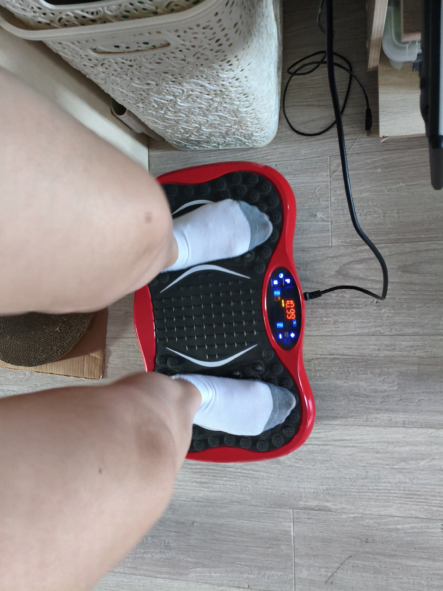 In this top-down shot, a person is standing on a red and black vibrating exercise platform. The person is wearing white socks. The platform has a digital display that shows "0.99" in orange digits, and several blue indicator lights. A black power cord is plugged into the side of the platform. The platform rests on a light-colored wooden floor. To the left of the platform, a portion of a tan-colored cardboard cat scratching post is visible. In the background, a white woven storage basket is seen on top of a wooden shelf unit. The setting appears to be indoors, likely a home, given the flooring and household items. The time of day is not discernible from the image. There are no overt emotional cues present. The only visible text is the numerical display on the exercise platform. The location context of Orenburg, Russia, is provided but not visually evident in the image.