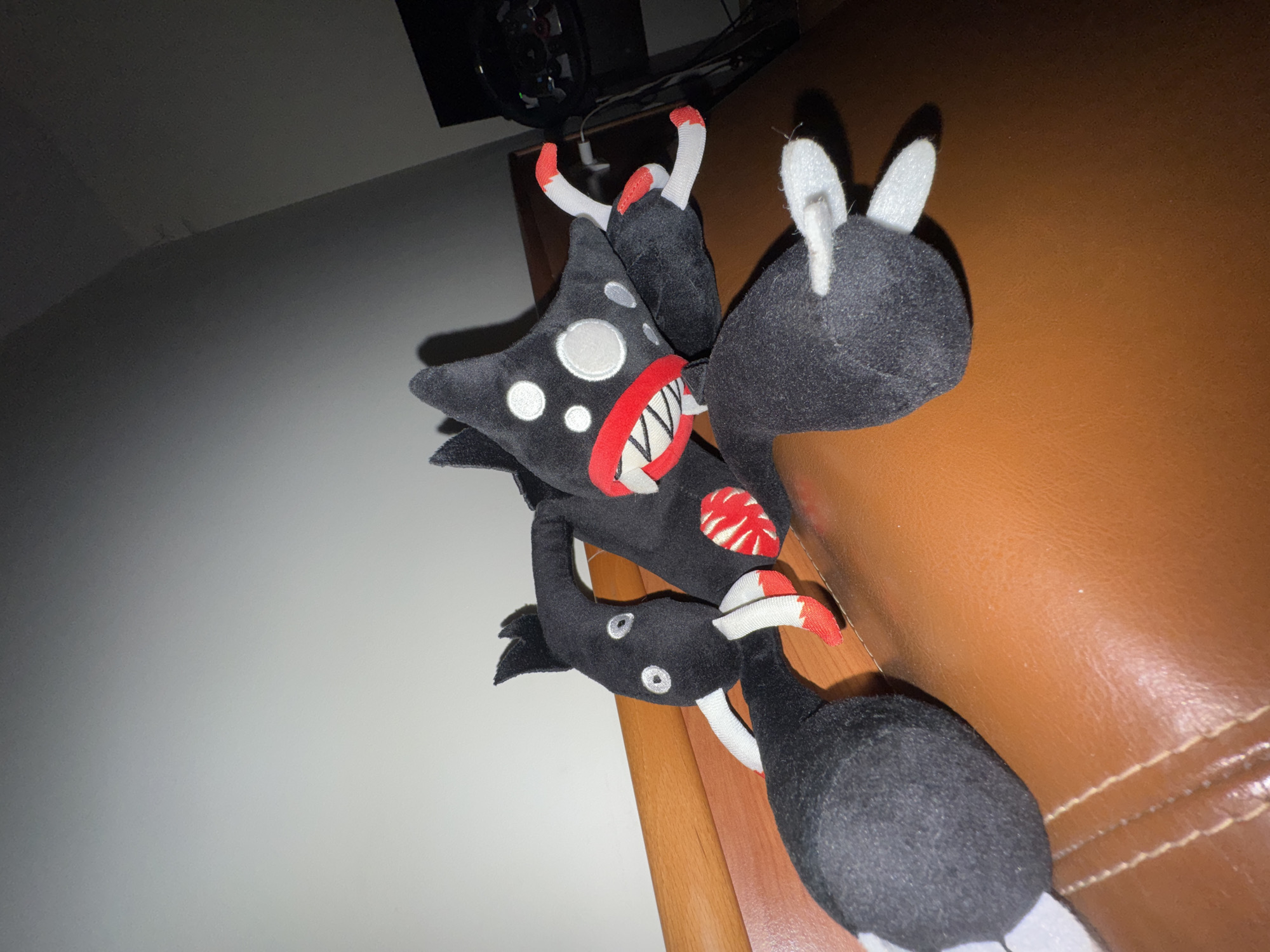 A close-up shot features a cluster of plush toys on a brown surface, possibly a leather couch or chair. The toys are predominantly black with red and white accents. The main toy has a star-like shape, with several white dots, a red trim around the mouth, and white pointed teeth. Its arms or appendages end in red and white striped segments. Another plush toy is attached and features white bunny ears. A third, smaller plush toy is partially visible. The setting is indoors, as suggested by the wall in the background. It is impossible to place this scene within the city of Varna, Bulgaria, without additional information.