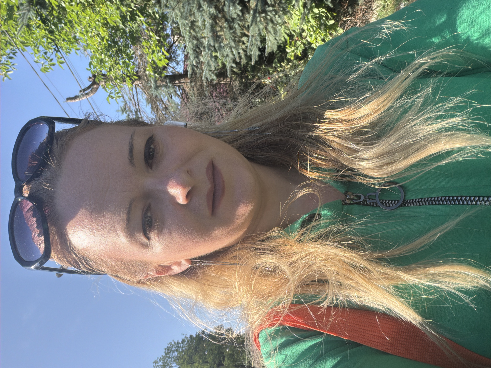 A close-up, outdoor, eye-level selfie shows a blonde woman with sunglasses perched on her head. She is wearing a green zip-up hoodie and an orange bag strap is visible over her shoulder. She has on long dangling earrings and white earbuds. The background consists of a clear blue sky, green trees, and power lines. The lighting suggests it is daytime, with the sun casting shadows and highlighting the subject's hair.