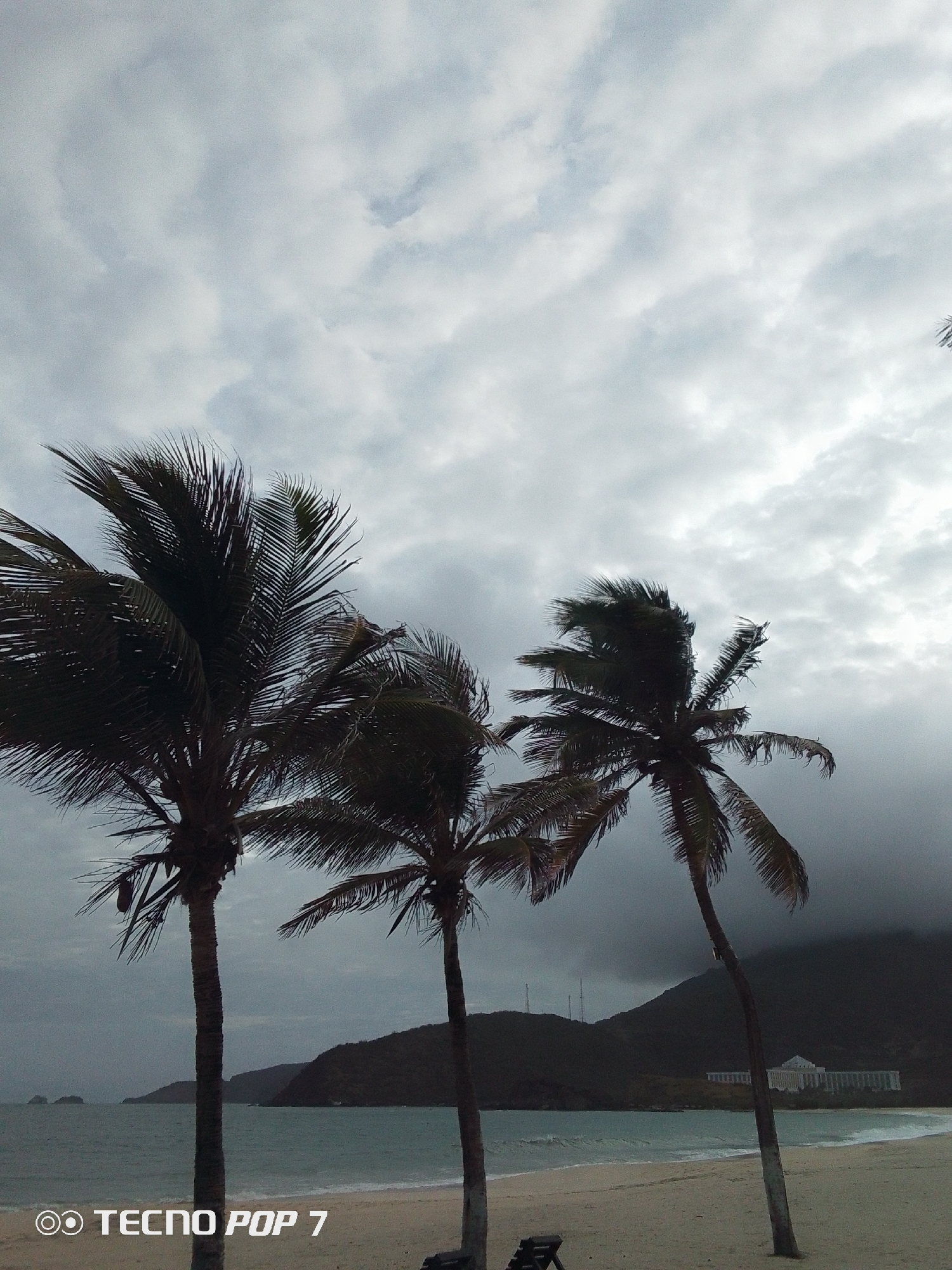The image captures a coastal landscape in Pedro González, Venezuela, under a heavily overcast sky dominated by dense grey and white clouds. Three prominent palm trees with dark, wind-swept fronds are silhouetted in the foreground on a light-colored sandy beach. The ocean, appearing turquoise-blue, features gentle waves breaking at the shoreline. In the mid-ground, a chain of dark, vegetated mountains and hills extends along the coast, with smaller islands visible on the left horizon. On the far right, a large, multi-story white building, consistent with a resort or hotel, stands at the base of a dark, cloud-shrouded mountain. Faint communication towers are discernible on a distant mountain ridge. A partial dark object, possibly a beach chair, is visible on the sand at the bottom center. No individuals are present.