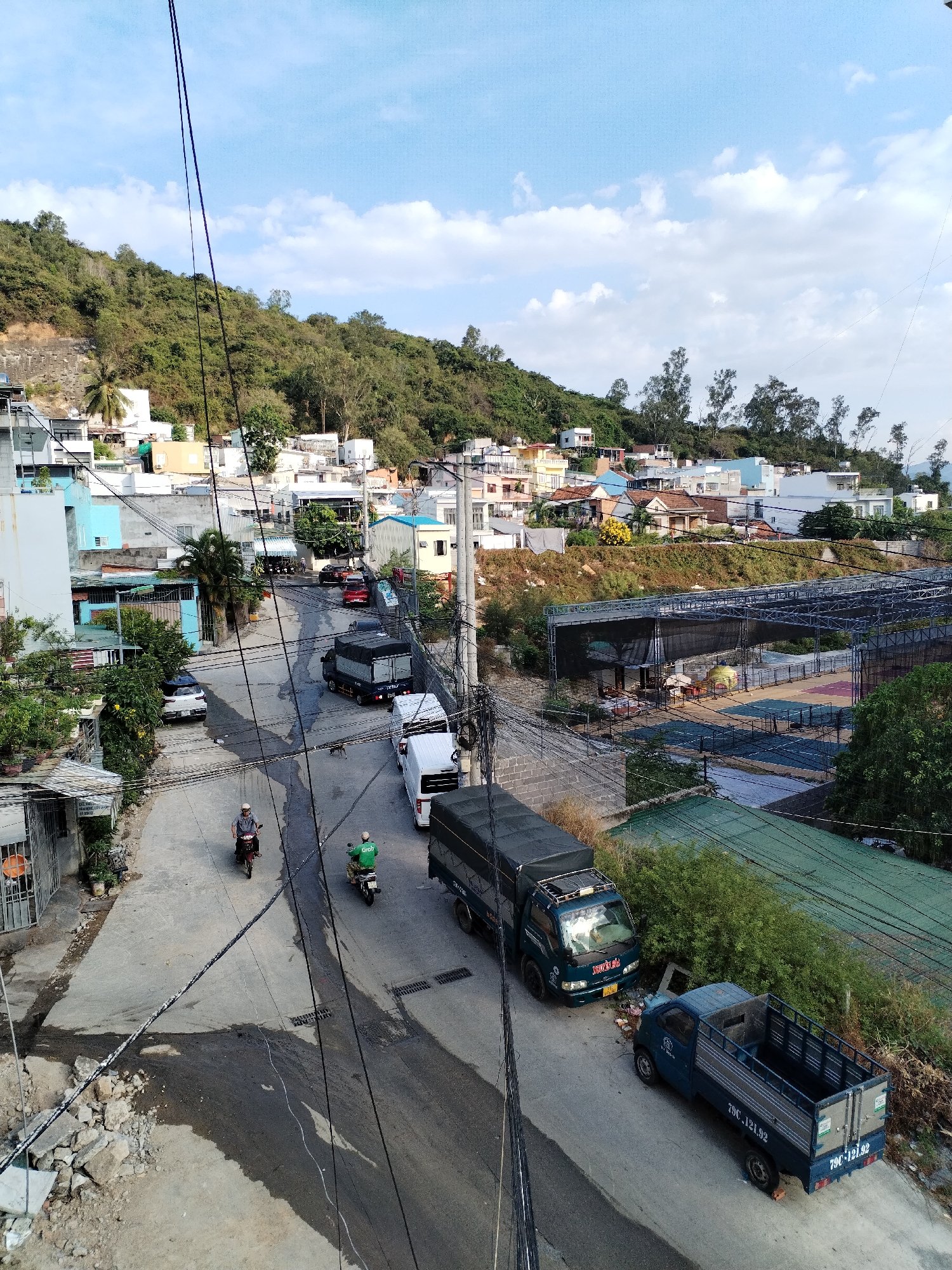 The image captures a street scene in the Nha Trang Ward area of Nha Trang, Vietnam. The view is elevated, providing a panorama of a residential area nestled at the base of a forested hill. The sky is partly cloudy.
The street below is active with vehicular traffic, including several trucks of varying sizes. A blue truck is parked to the side of the road. Two motorcycles are moving on the street, one ridden by a person in a helmet and the other with a person wearing a green shirt.
Buildings of different colors and styles are situated on the hillside. An open-air structure with a metal frame and covered area is also visible. Power lines crisscross the scene, adding to the visual clutter. Along the bottom left is a pile of rocks and debris.