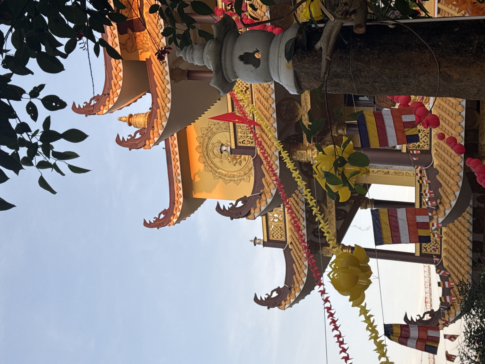 The image captures the ornate roofline of a Buddhist temple in Da Lat, Vietnam, under a clear sky. The temple features traditional tiered roofs with curved eaves, adorned with decorative elements and colorful flags. Yellow paper lanterns and strings of red and yellow pinwheels are strung across the foreground, adding a festive atmosphere. A stone lantern stands prominently in the right foreground, partially obscured by branches and leaves from a nearby tree. Several Buddhist flags, displaying horizontal stripes of yellow, red, blue, and white, are visible, some hanging from poles and others strung between buildings. The architectural style suggests a Vietnamese Buddhist temple, likely decorated for a festival or special occasion.