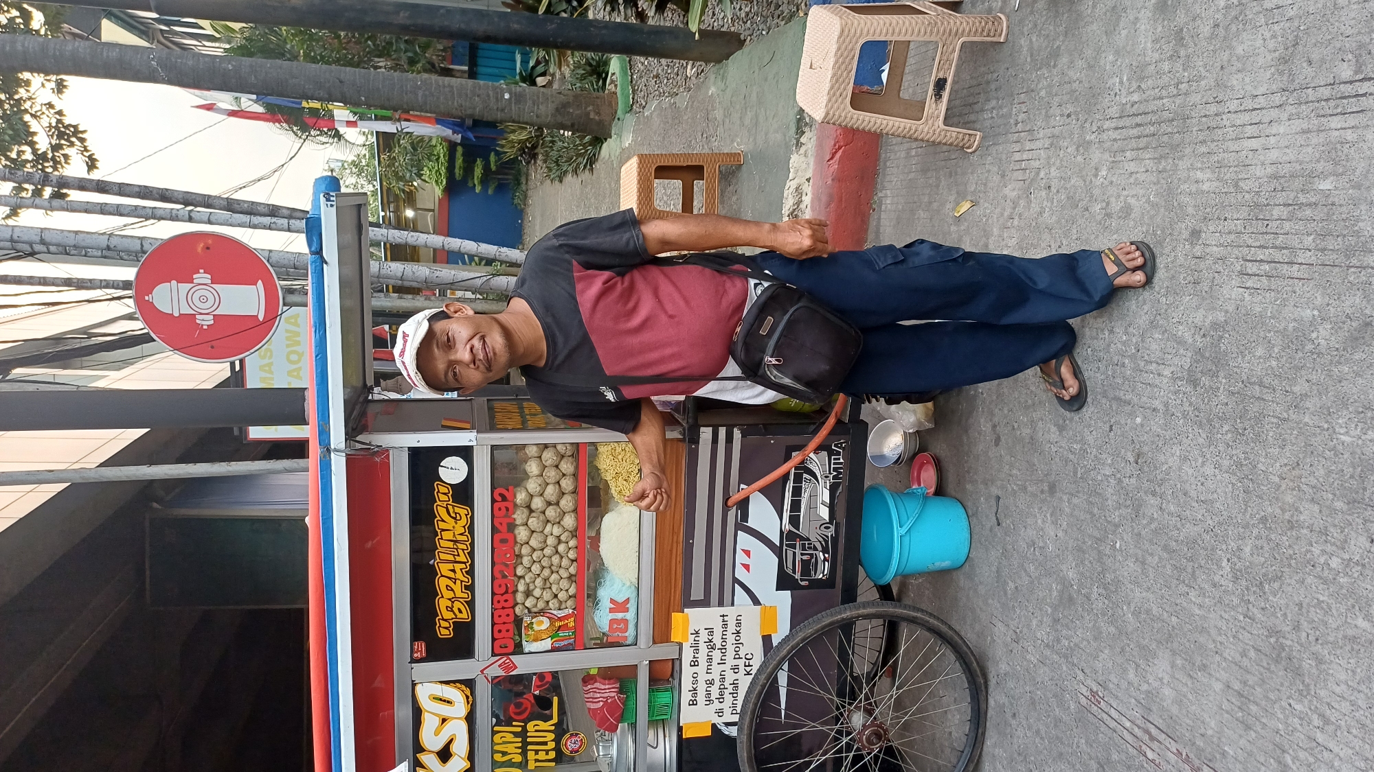 A middle-aged Indonesian man stands in front of a food cart on a concrete sidewalk in front of a building. The man is wearing a black, red, and white t-shirt, dark blue cargo pants, and black flip-flops. He has a small black shoulder bag slung across his chest. He is smiling and giving a thumbs-up with his right hand.
The food cart is colorful with red, white, and blue trim. It has a display window filled with meatballs, noodles, and other food items. A phone number, "08889280492", is visible on the cart. A sign on the cart reads "Bakso Bralink yang mangkal di depan Indomart pindah di pojokan KFC", indicating the food cart is named "Bakso Bralink" and has moved from in front of an Indomart to a corner near a KFC.
Behind the man and the food cart, there are palm trees, a building with a glass facade, and a red and white fire hydrant sign. Two small, tan plastic stools are visible on the right side of the image. A blue bucket and some bowls are on the ground to the left of the food cart. The overall scene suggests an outdoor food stall operating in a busy urban area. The specific location of South Jakarta, Indonesia is indicated by the presence of an Indomart and KFC, which are common retail chains in the region.