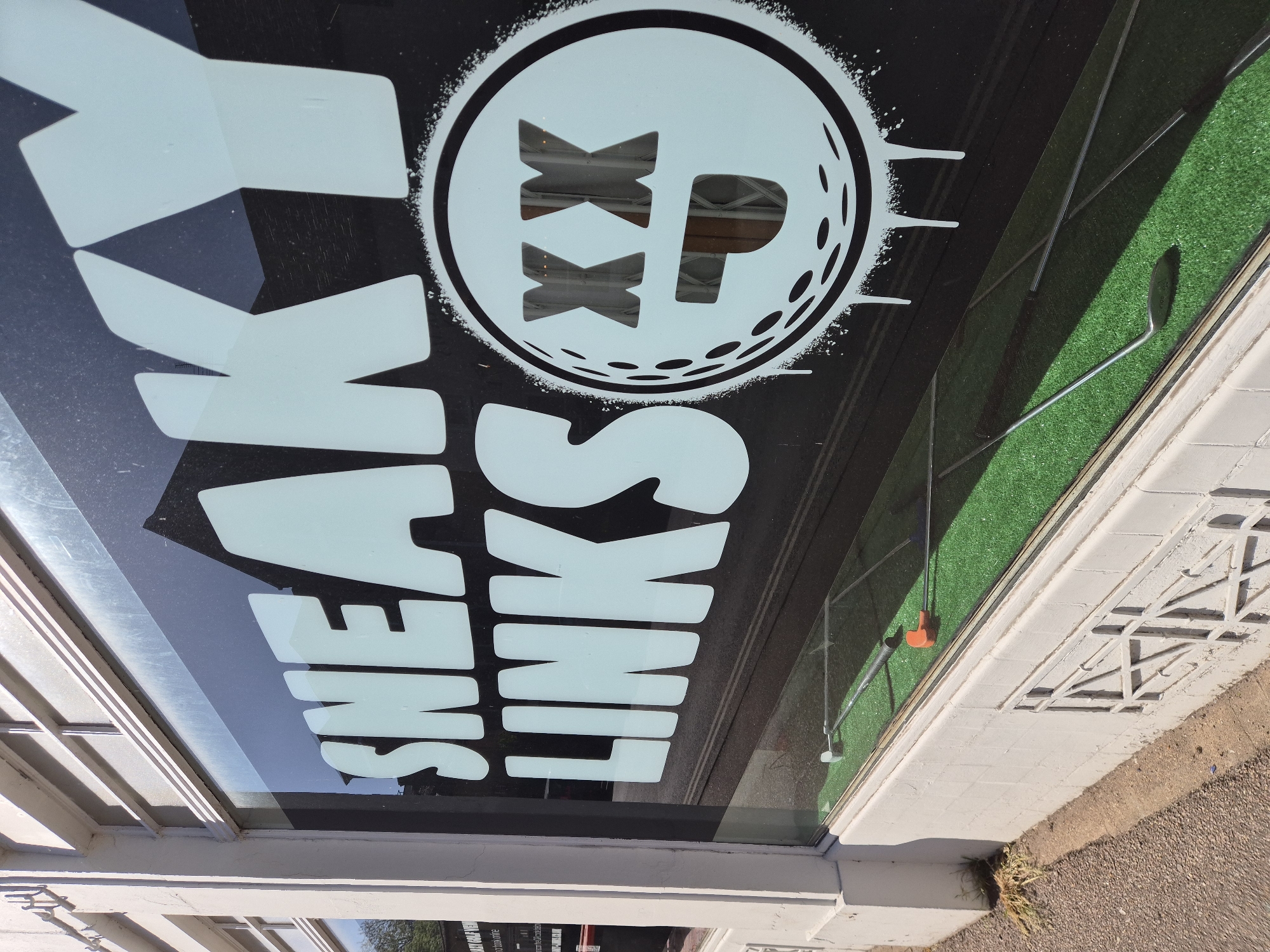 An outdoor, rotated shot captures the storefront window of a business, likely located in West Suffolk, United Kingdom, under bright daylight. The window features white vinyl lettering and a logo, suggesting a golf-related establishment.
The most prominent elements are the large white letters on the dark window, which, when rotated to the correct orientation, clearly spell "SNEAKY LINKS". To the right of the text, a circular logo is displayed. This logo depicts a stylized golf ball texture, within which are the letters "XX P". The entire logo has a "spray paint" or "distressed" outline.
Through the clear parts of the window, an interior scene is visible. A patch of green artificial turf is laid out, upon which a silver golf putter rests, its clubhead near an orange golf ball. This setup suggests an indoor mini-golf course, golf simulator, or practice area.
Reflections on the window show elements of the exterior environment, including parts of a building or canopy structure with white linear frames, and faint glimpses of distant trees against a bright sky. The bottom portion of the image shows a paved sidewalk or pathway next to the building. This pavement features decorative geometric patterns and some small weeds growing at the base of the wall, further grounding the scene in an urban or suburban setting.
