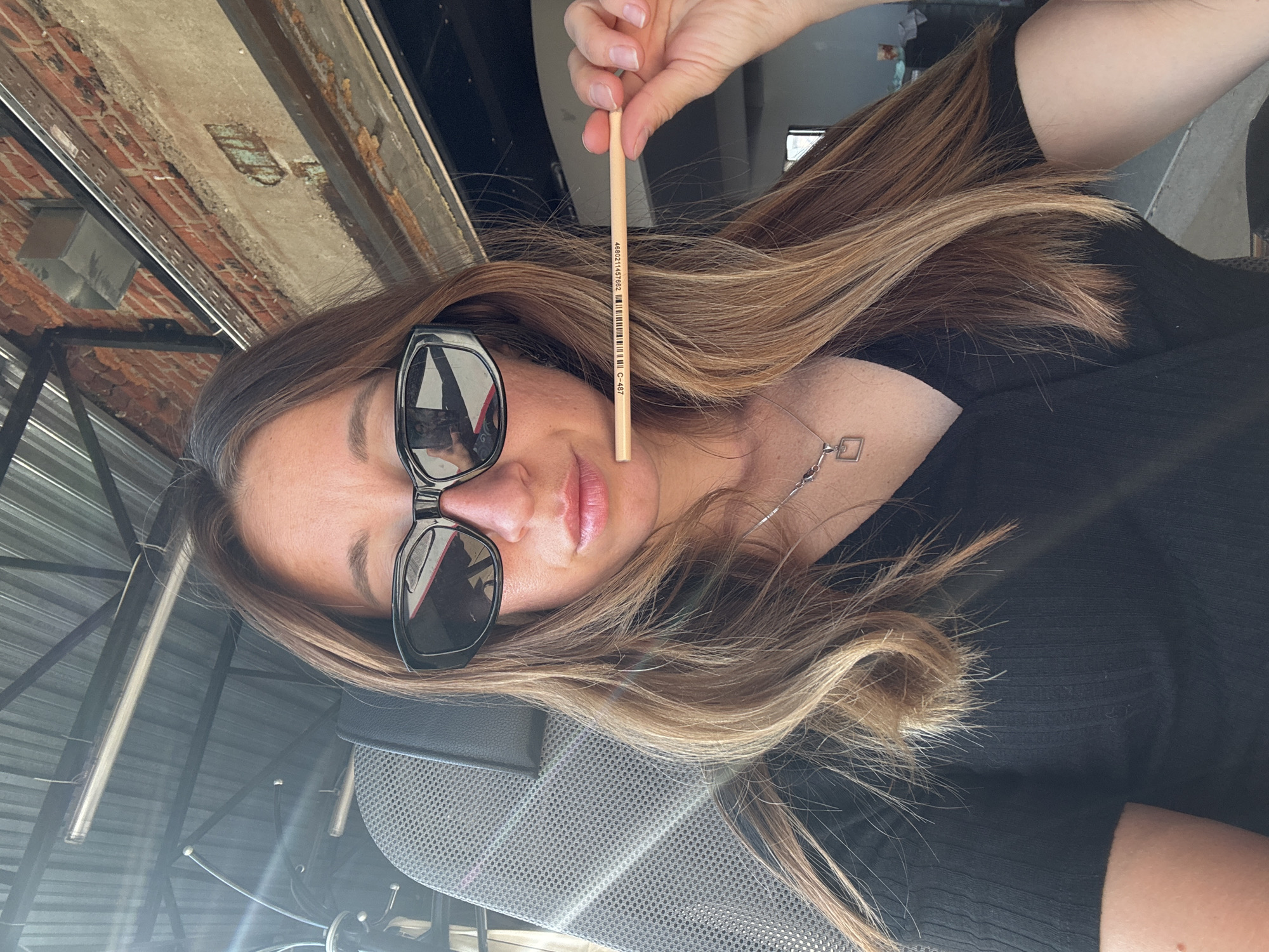A woman is holding a pencil in front of her face while seated. She wears sunglasses and a black top. The environment appears to be an urban setting with brick walls and metal structures visible. The location is claimed to be Chaikovsky, Russia, although this cannot be verified from the image.