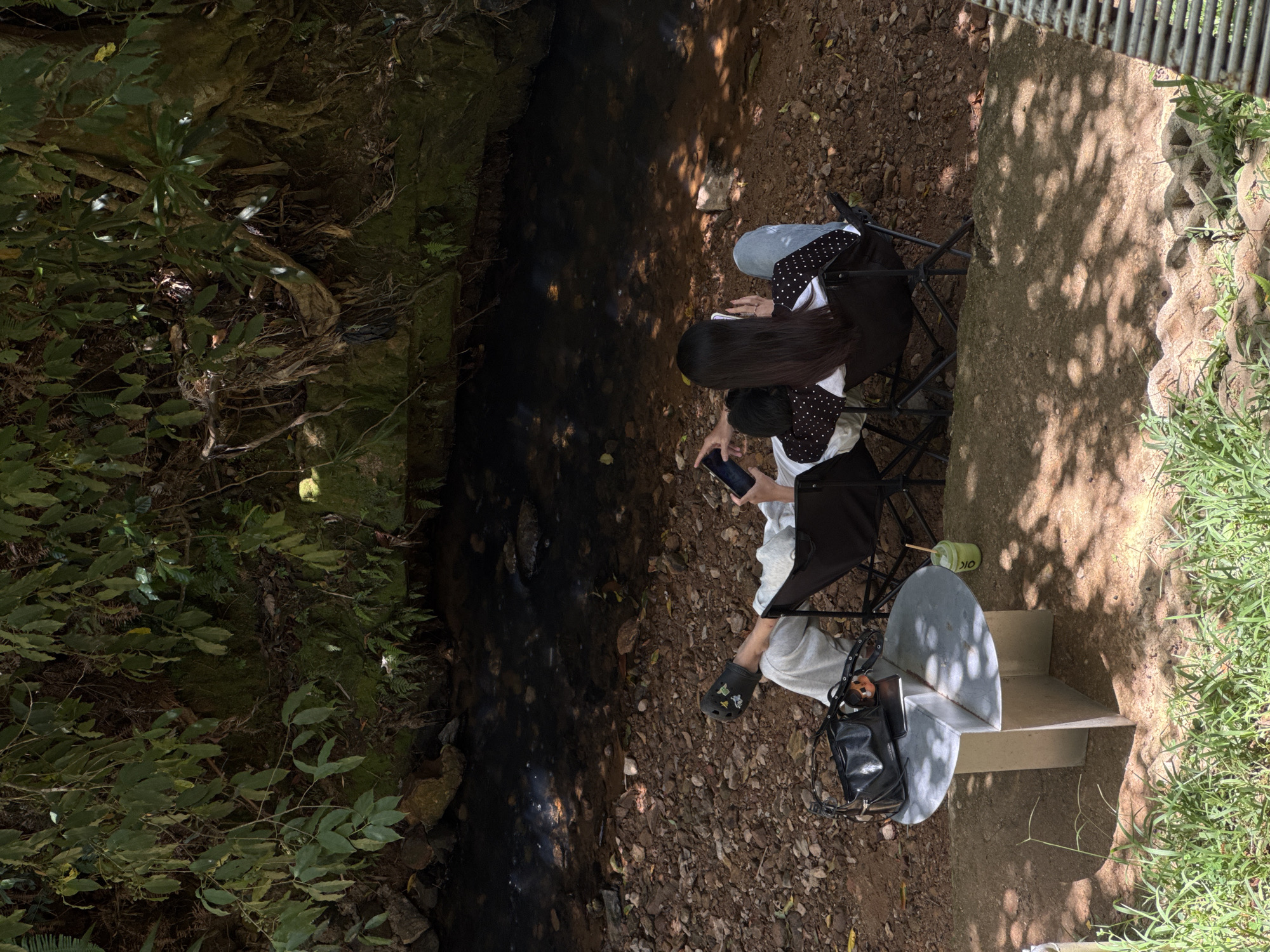 Two individuals are seated in camping chairs by a stream in Da Lat, Vietnam. The person on the left, wearing gray sweatpants and white sneakers with charms, is holding a smartphone. The person on the right, wearing blue jeans and a black and white polka dot shirt, is also holding a smartphone and has their arm around the other person's shoulder. A black handbag and a small, metal, four-legged table with a curved top are to the left of the chairs. A green cup with a straw is on the table. The stream flows through a rocky, wooded area with lush green foliage and mossy rocks along its banks. Sunlight filters through the trees, creating dappled shadows on the ground.