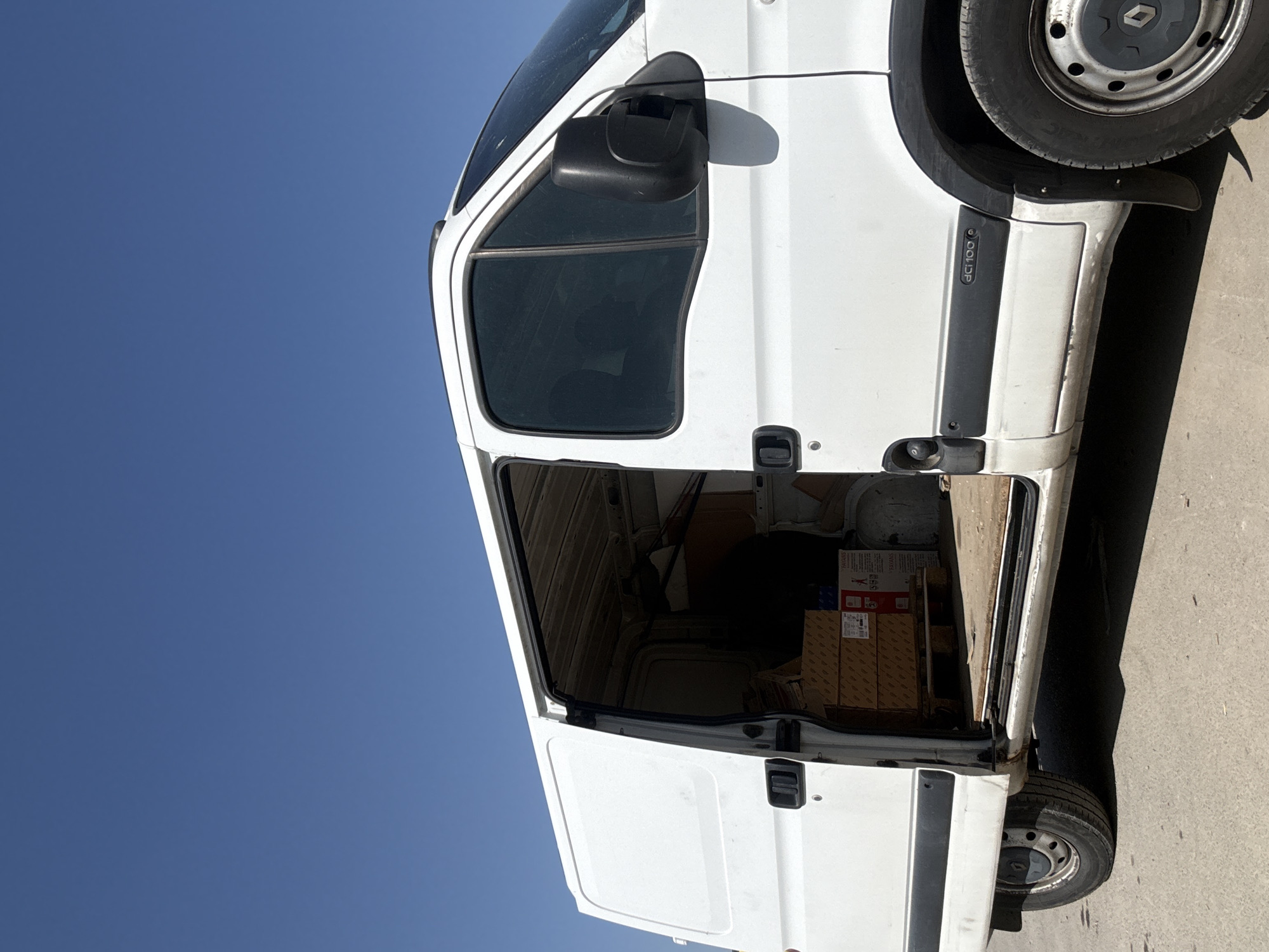The image displays a white commercial van, likely a cargo or delivery van, parked outdoors under a clear, bright blue sky. The photograph is oriented vertically, showing the side of the van with both the front passenger door and the side sliding cargo door wide open.
Inside the van's cargo compartment, several cardboard boxes are visible, suggesting the vehicle is used for transporting goods. Text partially visible on some boxes includes "HAVOLINE" and what appears to be "TAXANS". The interior of the cargo area shows functional paneling.
The ground surface beneath and around the van appears to be paved, possibly concrete or asphalt, with distinct shadows indicating bright sunlight and a clear day, likely during the mid-day or afternoon.
Notable features on the van include a black side mirror on the passenger door and black protective trim along its side. Above the track for the sliding door, the text "dCi 100" is clearly visible, often denoting engine specifications. The front wheel on the visible side features a dark hubcap with the recognizable Renault logo.
There are no people present in the image, and no specific activity such as loading or unloading is actively depicted, though the open doors imply readiness for such tasks or a recent stop. The scene captures a functional vehicle in an outdoor, sunny setting, consistent with a location like Busmantsi, Bulgaria.