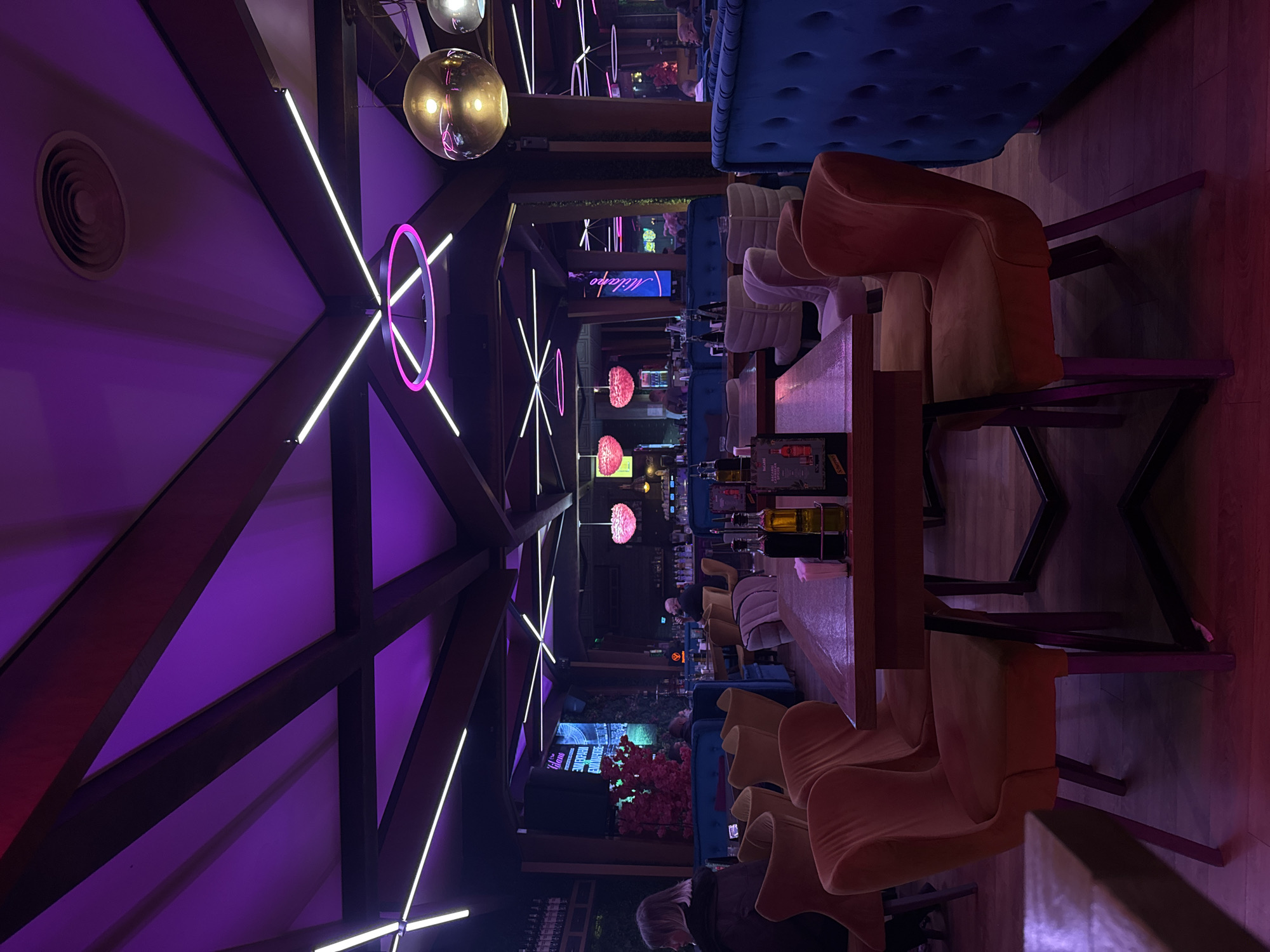 The image shows the interior of a restaurant or bar in Sofia, Bulgaria, based on the "Milano" sign visible. The scene is lit with purple and pink neon lights. The room has a high, vaulted ceiling with exposed wooden beams and decorative lighting fixtures. There are tables and chairs set up, with some tables having bottles and glasses. The seating includes blue upholstered booths and light-colored chairs with curved backs. A few people are visible in the background, sitting at the bar and at other tables. Greenery, including plants and floral arrangements, is present. The focus of the photograph is the rows of tables and chairs leading into the depth of the restaurant.
