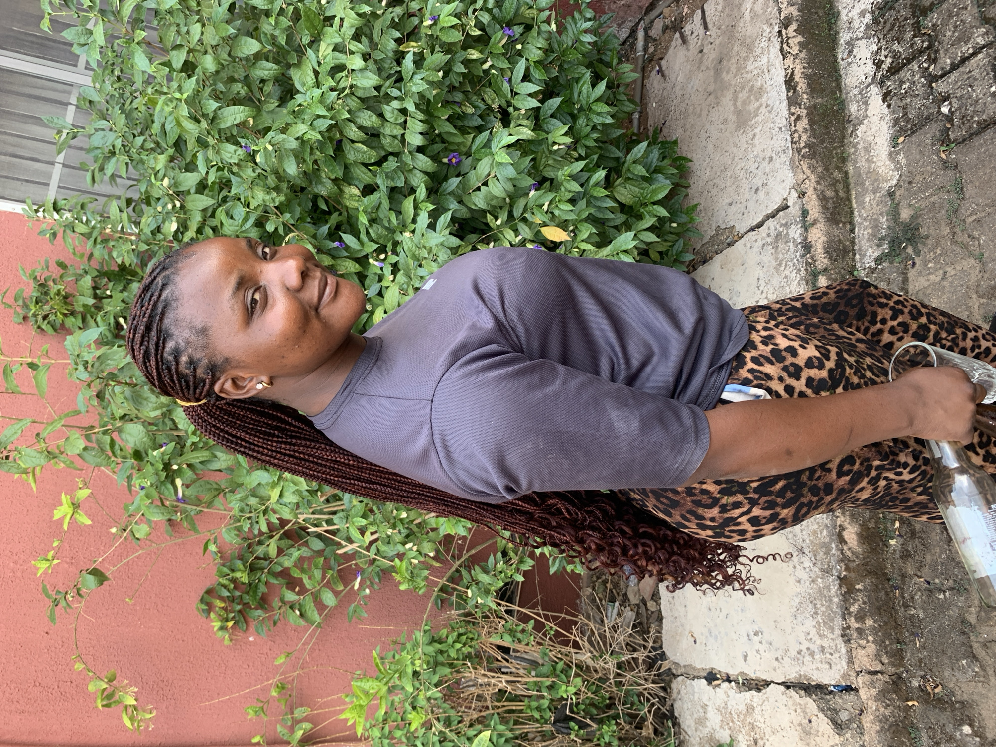 An outdoor, vertically oriented full-body portrait features a young Black woman standing or leaning, holding a clear glass bottle in her right hand. She has long, dark brown braided hair, with the lower sections styled into curls, and wears a light-colored stud earring. Her attire consists of a plain gray short-sleeved top and distinct leopard-print leggings or pants. Her head is tilted slightly, and she looks towards the camera with a calm, faint smile.
The setting appears to be a daytime outdoor location, likely in Jos, Nigeria, given the context. Behind the woman is a dense, vibrant green bush adorned with small purple flowers, contrasting with a reddish-brown textured wall on the left side of the frame. The ground beneath her is a cracked concrete pathway, showing signs of wear and some sparse, dried vegetation. The clear glass bottle she holds seems mostly empty, with some liquid residue and an indistinct white label partially visible near her hand.