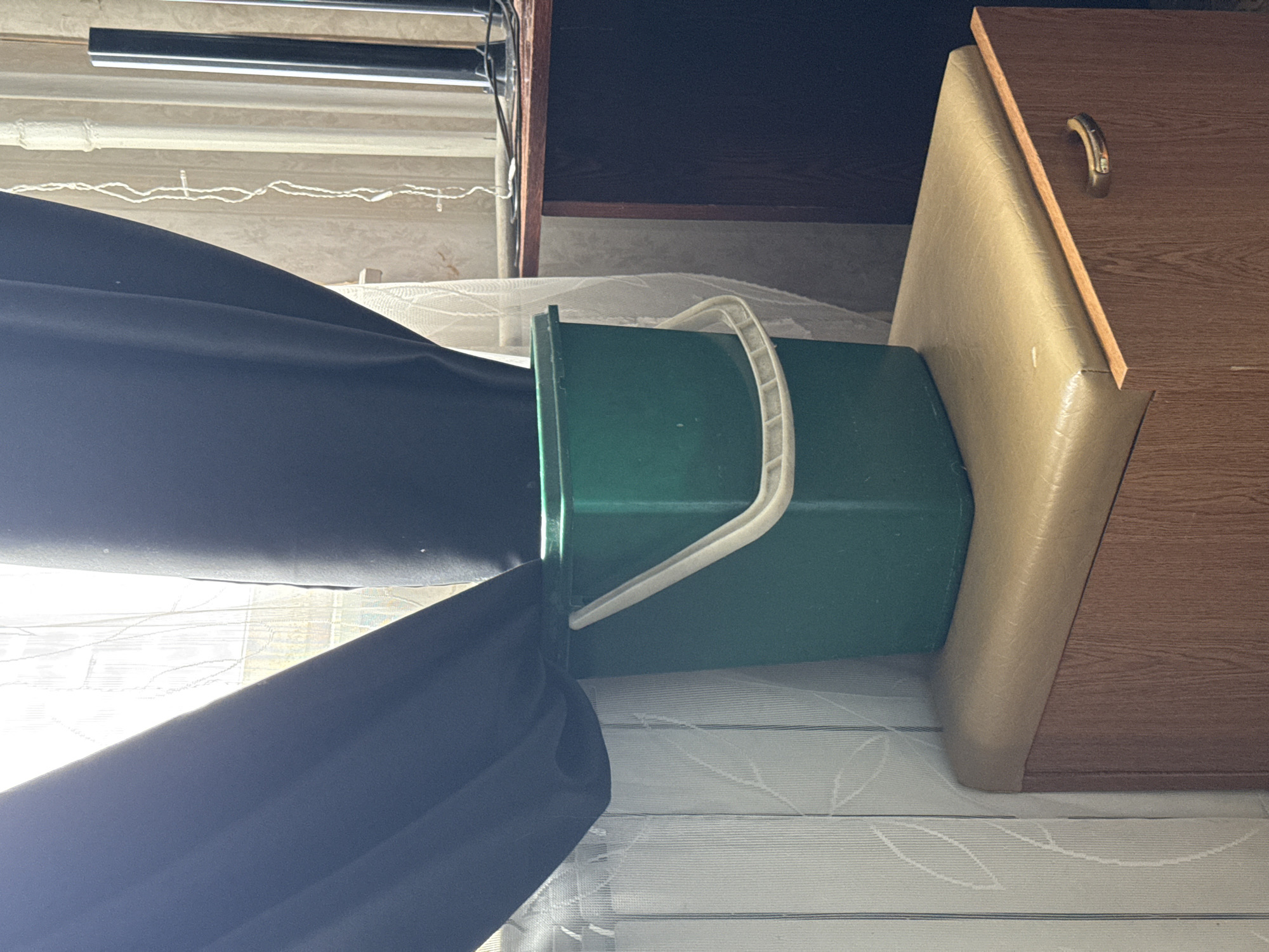 A green plastic bucket with a white handle sits next to a wooden cabinet on top of a white patterned surface. A black curtain is draped across the bucket. The location is not discernable from the image, thus the request to include the city of Khimki, Russia, is not possible to accommodate.