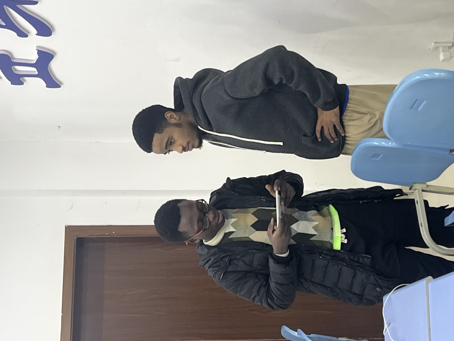 The image displays an indoor scene in Jiangjin, China, featuring two adult males of dark complexion. On the left, a male with short dark hair, wearing red-framed glasses, a black puffer jacket over a cream, brown, and dark green patterned sweater, and black trousers with a visible neon yellow-green elastic band, holds a white rectangular electronic device horizontally with both hands, intently looking down at its screen. To his right, a second male with short dark hair and faint facial hair, dressed in a dark grey hooded sweatshirt with a white drawstring and light brown pants, stands with his hands on his hips, facing left and observing the first male and the device. The background includes white walls, a dark brown wooden door on the left, and partially visible blue Chinese characters on the upper right wall. In the foreground, several light blue plastic chairs with metal frames and a light blue desk surface are visible.