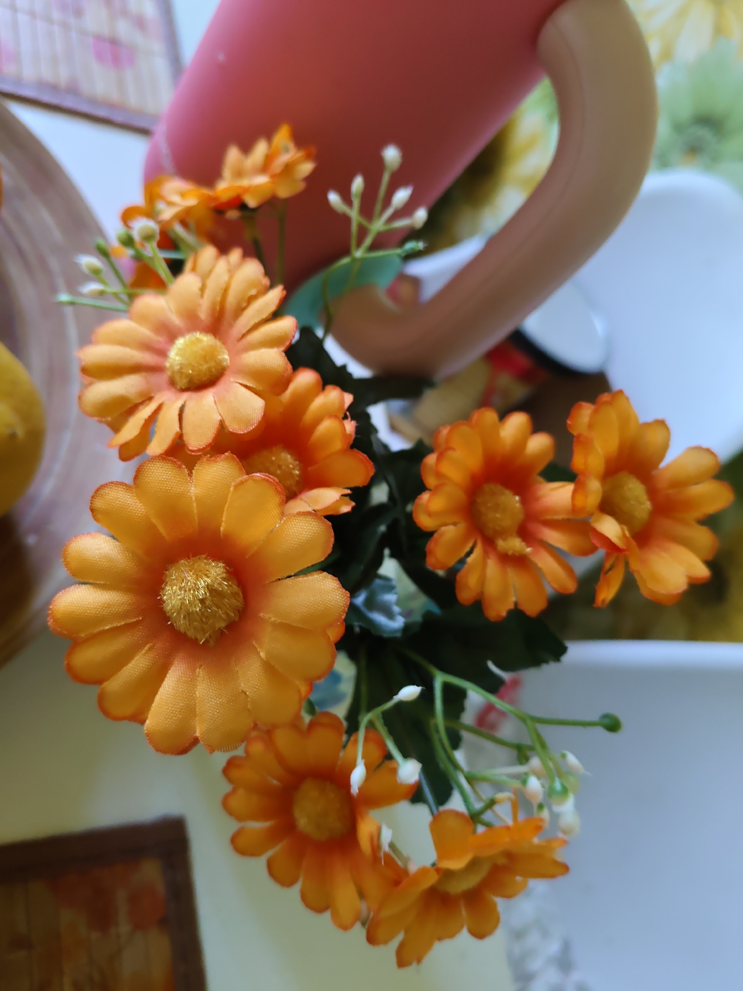 The image is a close-up shot of a bouquet of artificial orange daisies with yellow centers. The flowers are clustered together and appear to be part of a larger arrangement or decoration. Some green leaves are visible among the flowers. In the background, out of focus, there is a pink object that resembles a watering can or a pitcher, and part of a white object, possibly a mug or bowl. There is also a hint of a framed picture on the lower left. The scene appears to be indoors. The image does not provide any specific visual cues that would allow for the determination of its location in the city of Rostov-on-Don, Russia.