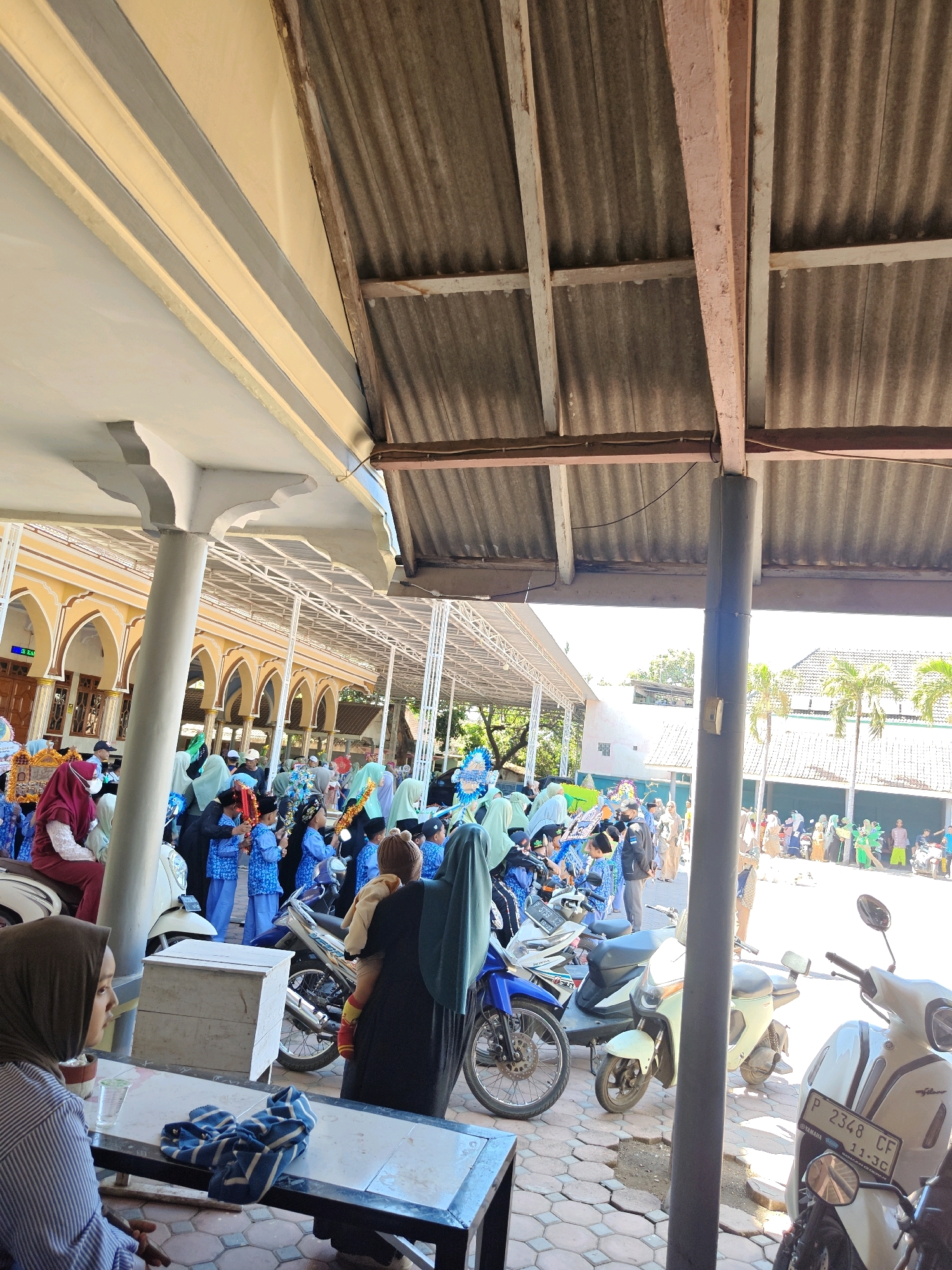 The image shows an outdoor scene, likely in Klampokan, Indonesia, with a crowd of people gathered. In the foreground, a woman wearing a striped long-sleeved shirt and a headscarf is seated at a table. On the table are a glass of water and a blue and white striped cloth. Behind her, a woman in a dark outfit, also with a headscarf, is holding a child. Several motorcycles are parked in the area.
In the background, a large group of people, many of whom appear to be children in blue outfits and headscarves, are standing and moving. There are also adult figures, some in traditional attire. The architecture visible includes a building with arched windows and a yellow facade, suggesting it might be a mosque or a school. A corrugated metal roof structure is prominent in the upper portion of the image, supported by wooden beams and a dark cylindrical pillar in the foreground. The ground is paved with hexagonal tiles. Palm trees and other vegetation are visible in the distance. The overall atmosphere suggests a community gathering or event.