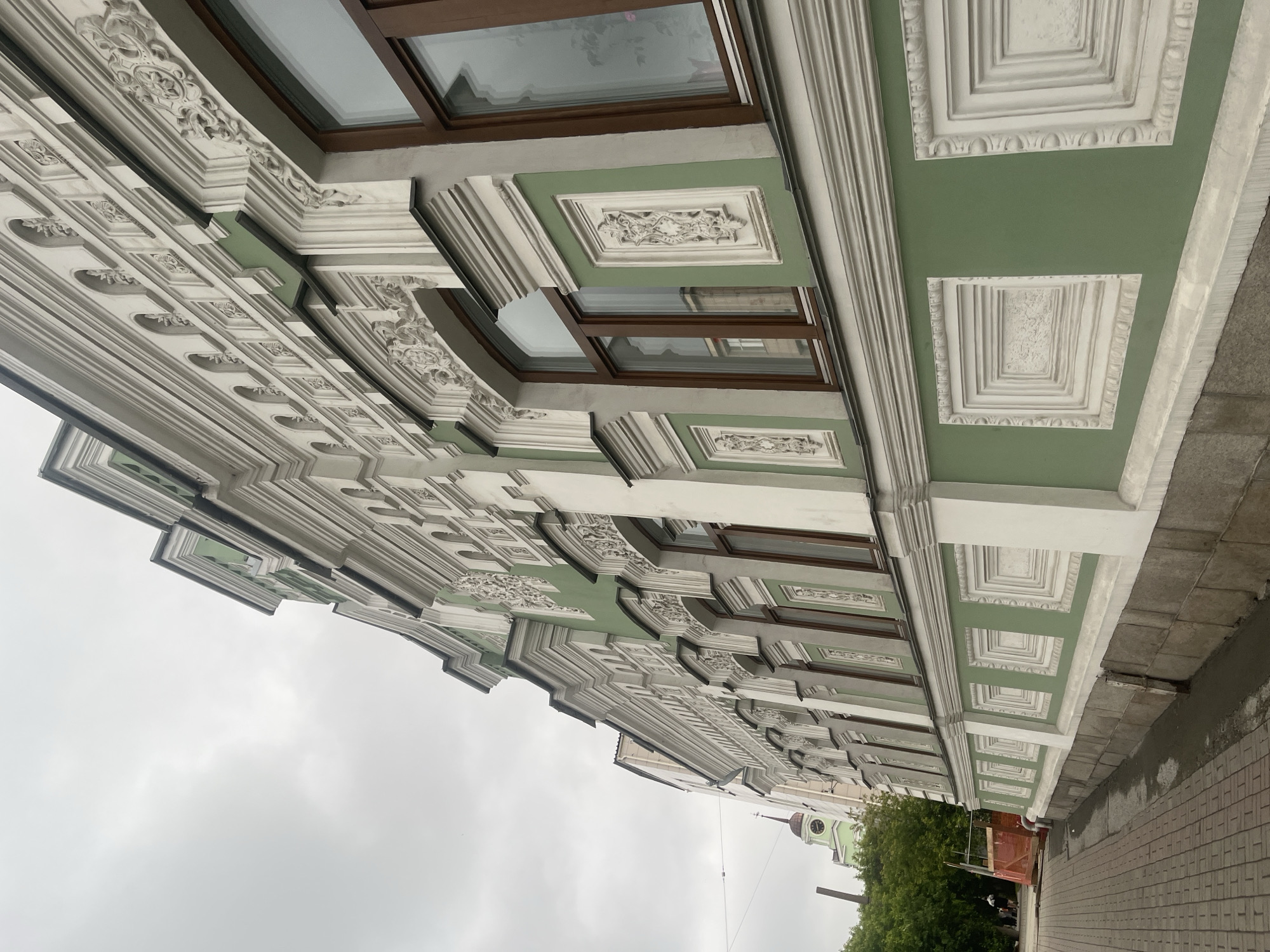 An eye-level, full shot captures the side of an ornate, multi-story building with a pale green facade. The building features elaborate white stucco detailing above the windows and along the cornices, with recessed panels and decorative moldings. The windows have dark brown frames. To the left, a brick sidewalk runs alongside the building, and further left, trees and a distant green tower with a clock are visible. The sky is overcast and gray.