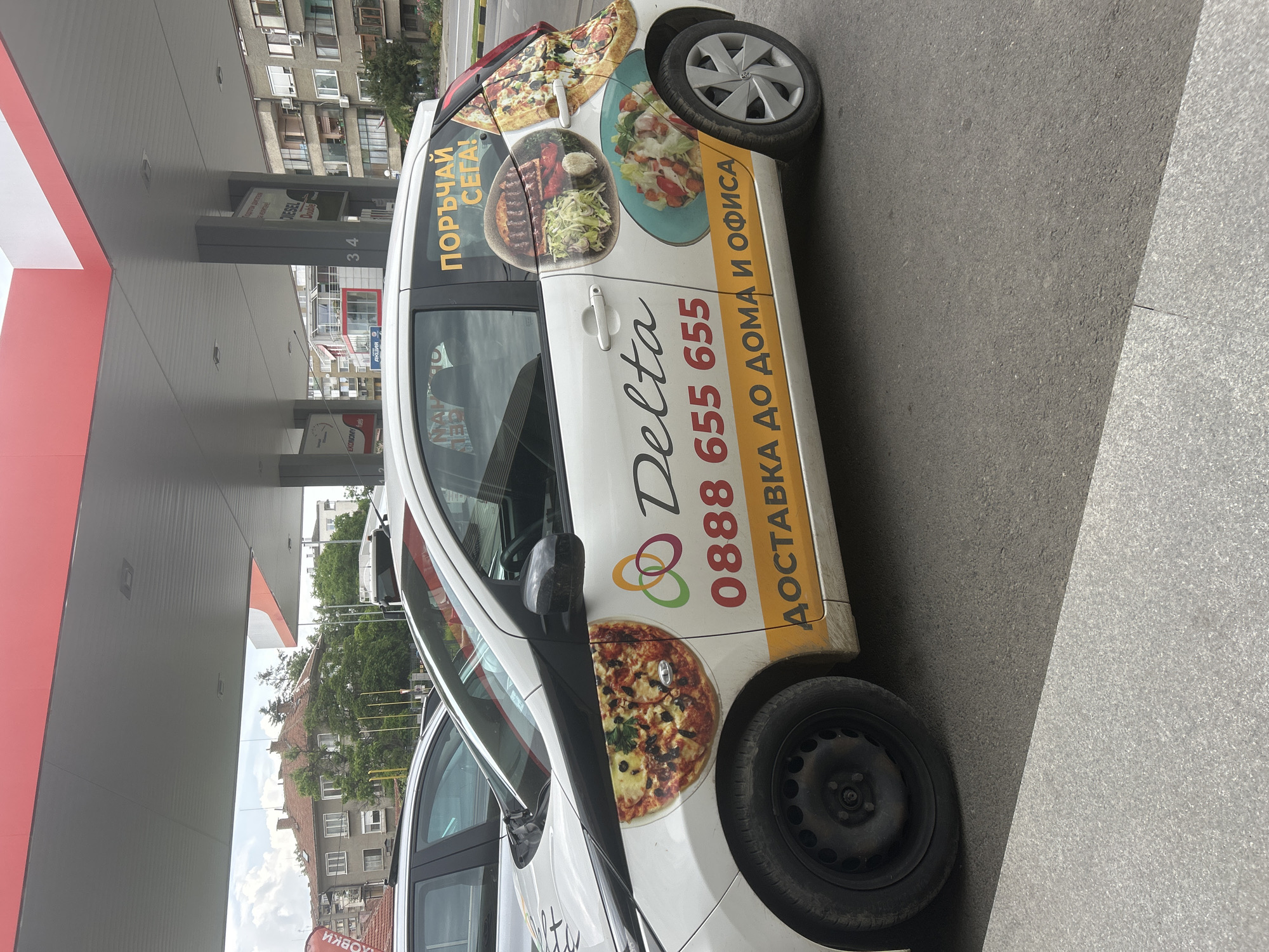 The image shows a white car parked next to a gas station. The car has "Delta" with contact information and images of food, including pizza, printed on its side, indicating it might be used for food delivery. In the background is the gas station, with its red and grey roof covering the fuel pumps, and buildings typical of an urban setting in Kazanlak, Bulgaria. Signs on the gas station pumps appear to say "DIESEL Double" and "EKONOMY fuels." The car is parked on a grey asphalt surface next to a grey tiled sidewalk.