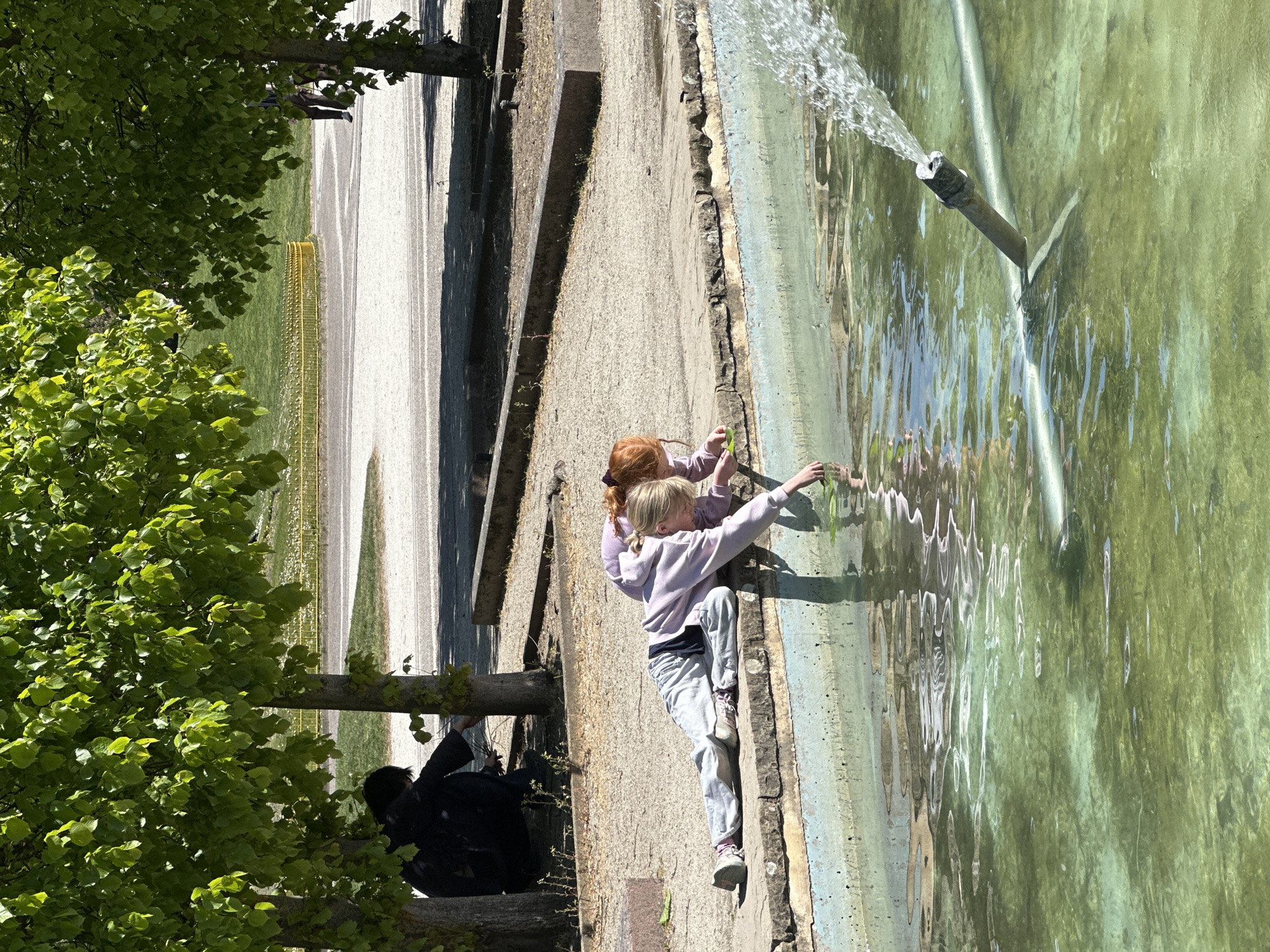 Two girls are standing at the edge of a long fountain. One of the girls has long blond hair and is wearing a lavender-colored hoodie and light gray pants. She is leaning slightly into the fountain and touching the water. The other girl has red hair and is standing behind the blond girl, also touching the water. Water is flowing from a metal pipe into the fountain. A stone path and trees line the side of the fountain. A yellow fence runs parallel to the path. A person in a black jacket is sitting on a tree stump. This is most likely in Kensington Gardens, London, United Kingdom.